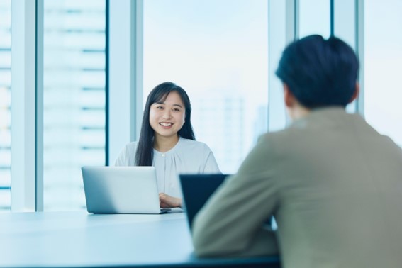 オフィスの会議室で向かい合って座る男女の写真。女性はノートパソコンを使い、男性は背中を向けている構図で、ビジネスの打ち合わせの場面を示している。