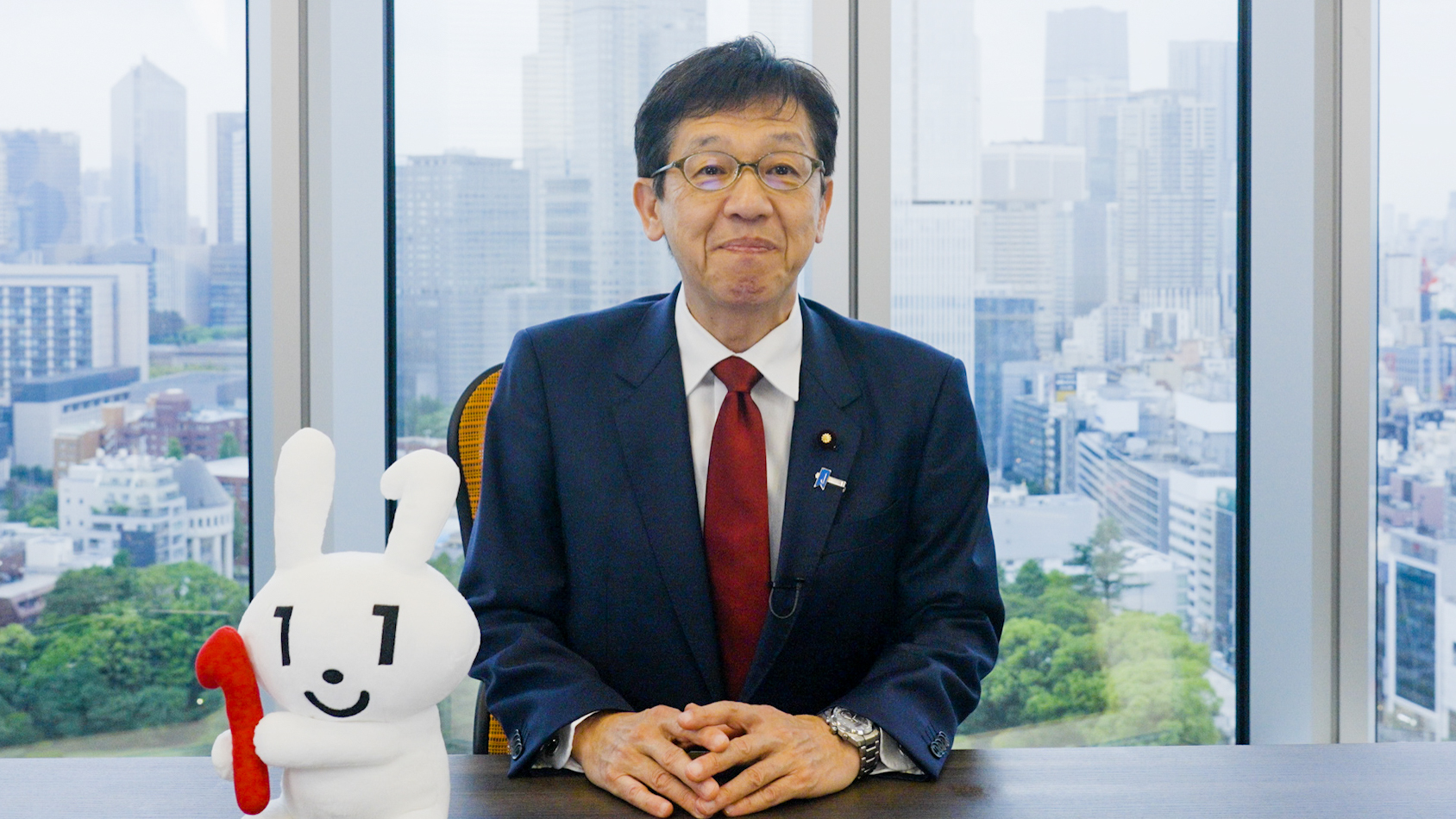 松本デジタル大臣が椅子に座り、デスクの上で手を組んでいる写真。デスクの上にはマイナちゃんのぬいぐるみも飾られている。