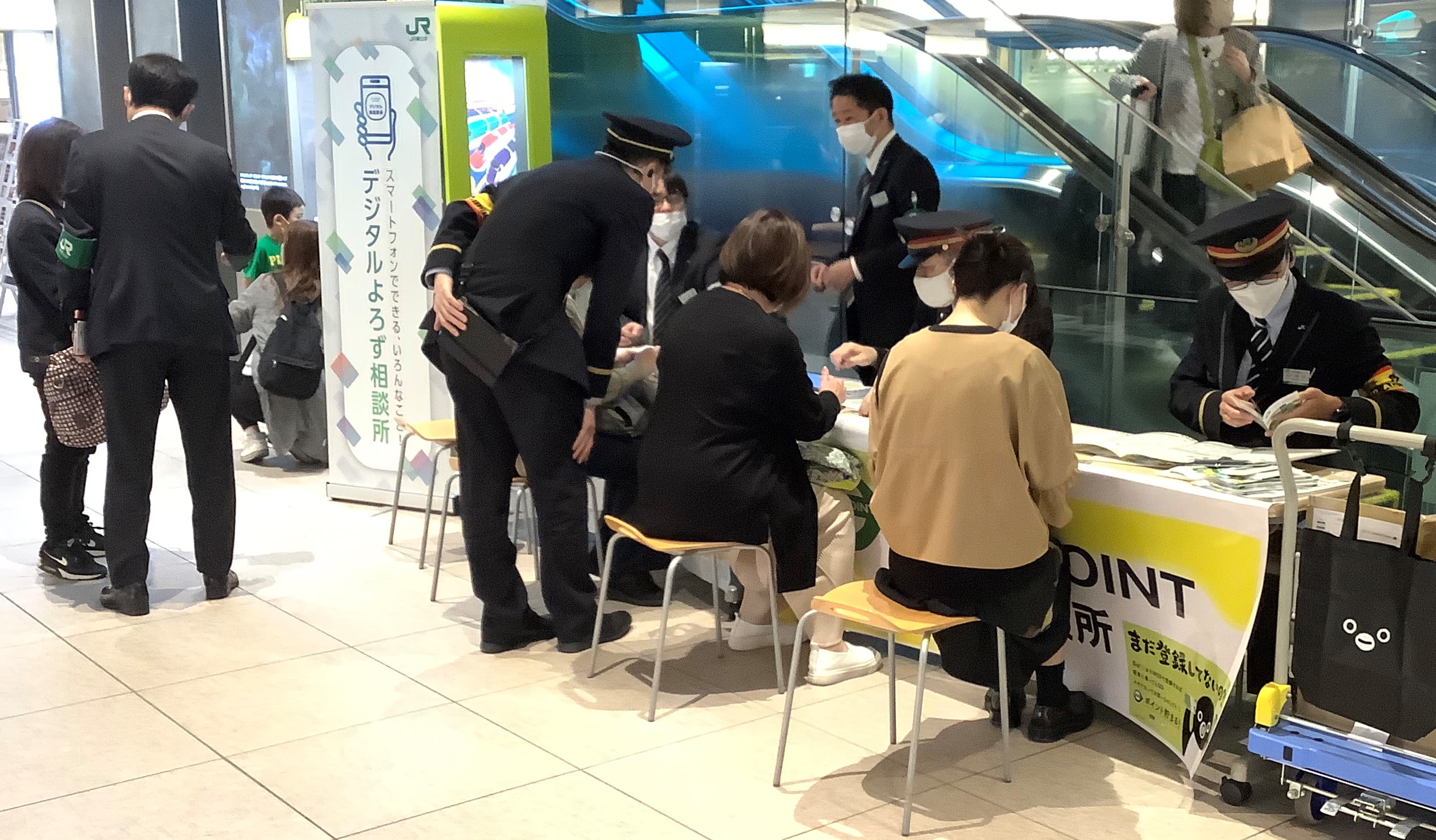 JR駅構内に設置されたデジタルよろず相談所の光景。垂れ幕右横に設置された大きめの長机にスタッフ二人がそれぞれが来訪者二人に個別に対応中。もう一人の来訪者には着席したスタッフ一人と立ったスタッフが対応中。さらに着帽したスタッフが立ったまま来訪者の手元を肩越しにのぞき込んでいる。