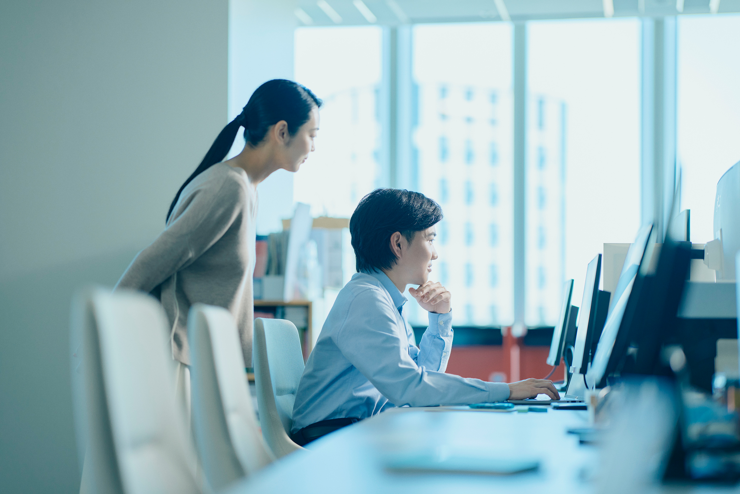 明るいオフィスの一角でモニタと椅子が並ぶデスクに座った男性と立っている女性が、一緒にパソコンの画面を見ている写真。男性は水色のシャツを着て椅子に座り、顎に手を当てて考え込むような表情でモニターを見つめている。女性はベージュのニットを着て、男性の左後ろに立ち、同じ画面を見下ろしている。背景には大きな窓から明るい自然光が差し込み、オフィスの棚やデスクが見える。全体に青白い色調である。