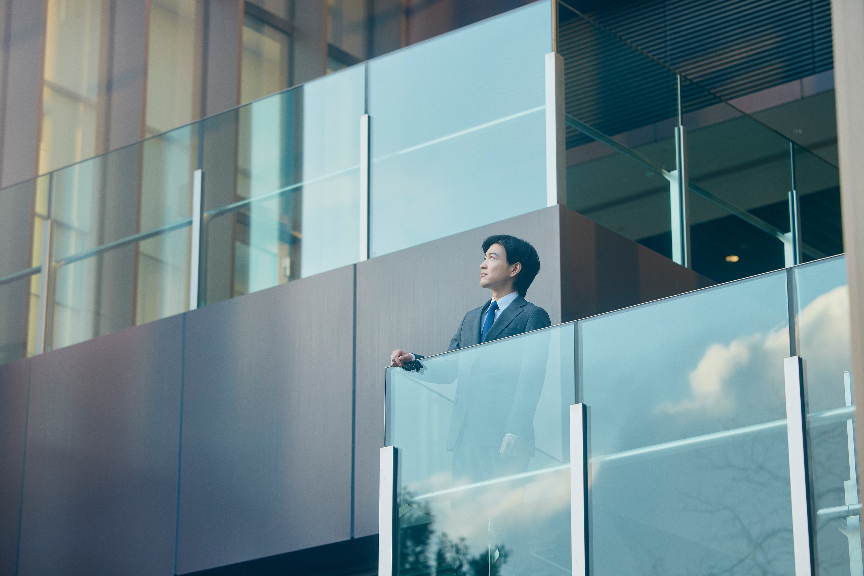 紺色のスーツにネクタイを着用した若い男性が、モダンなオフィスビルの中層階のガラス張りのバルコニーに立ち、ガラスの手すりに手を置き、斜め上方を見上げている写真。背景には現代的な建築のガラスと金属のファサードが広がり、ガラスには外の景色が映り込んでいる。全体に青みがかった落ち着いたトーン。