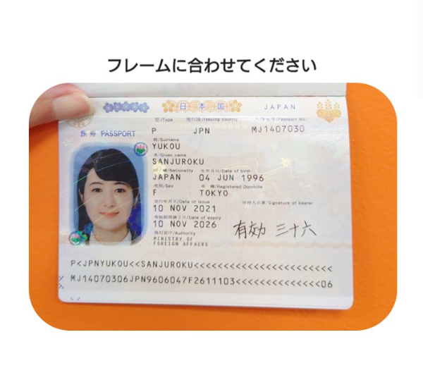 パスポートの写真。パスポートの顔写真が入っているページを指で開いてはっきり見えるよう机の上に置いている。写真の上には『フレームに合わせてください』という文章が入っている