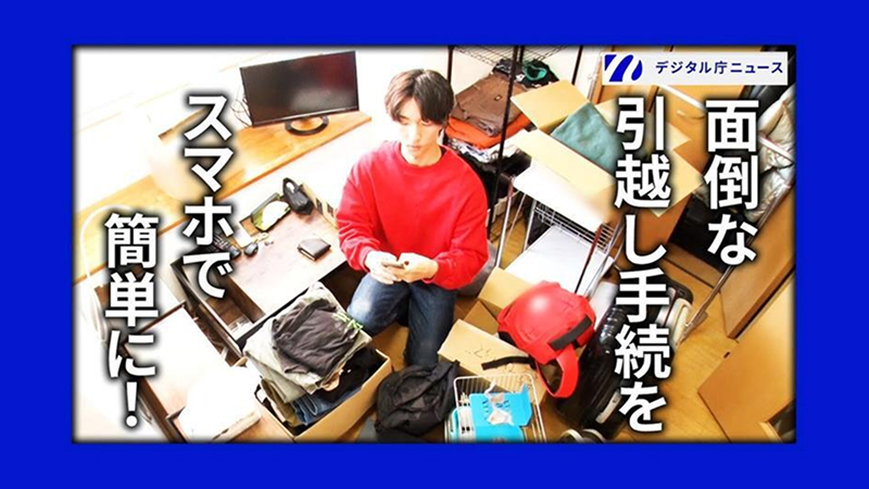 Digital Agency News' video thumbnail image. The logo of "Digital Agency News" in the upper right. "Complicated moving procedures" is written in white bold letters on the right side, and "Easy with your smartphone!" is written in large letters on the left side. The photo shows a man in a red sweatshirt sitting in the center of a moving room littered with cardboard boxes and luggage, using his smartphone from above. 