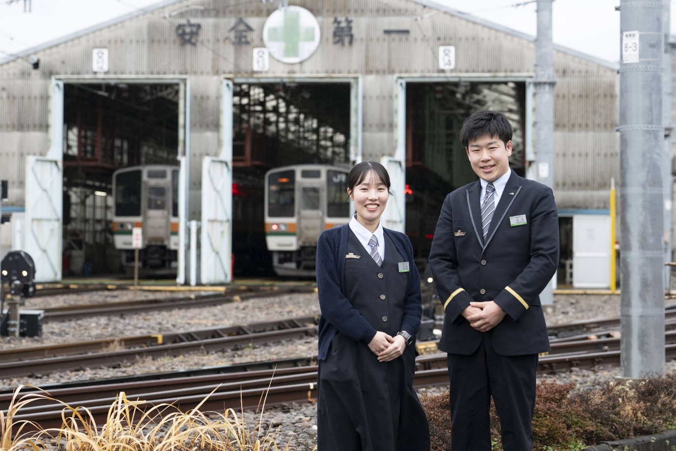 JR東日本の制服を着た20代と思われる女性社員と男性乗務員の2名が、車両基地の前に並んで笑顔を見せている。左側の女性はネイビーのカーディガン制服にストライプのネクタイと名札を着用。右側の男性はネイビーのスーツ制服に袖に金色のラインが入り、ストライプのネクタイと名札を着用。両者とも手を前で重ね、穏やかな笑顔で正面を向いている。背後には金属製の大型格納庫があり、建物正面に『安全第一』の文字と緑の十字マークが掲示されている。格納庫内にはJRのオレンジ・緑帯の車両が2編成格納されており、手前には線路が通っている。
