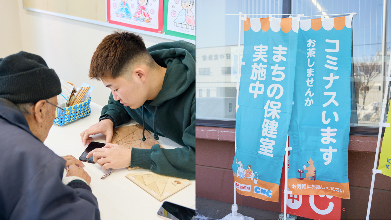 画像は左右ふたつに分かれている。左側は、室内のテーブルで、グレーのパーカーを着た若い男性（20代前後）が、黒いニット帽をかぶった高齢男性に向かって身を乗り出し、スマートフォンの画面を指し示しながら操作方法を説明している。テーブルの上には木製のパズル（タングラムのような三角形のピース）が置かれており、壁には子ども向けの絵や掲示物が貼られている。右側は、屋外に設置された2本の水色ののぼり旗。右側には「コミナスいます　お茶しませんか　お気軽にお越しください」、左側には「まちの保健室　実施中」と白い文字で縦書きされている。旗の下部には郵便局・CNCのロゴと、住民が集う様子のイラストが描かれている。オレンジ色の縁取りが施されている。