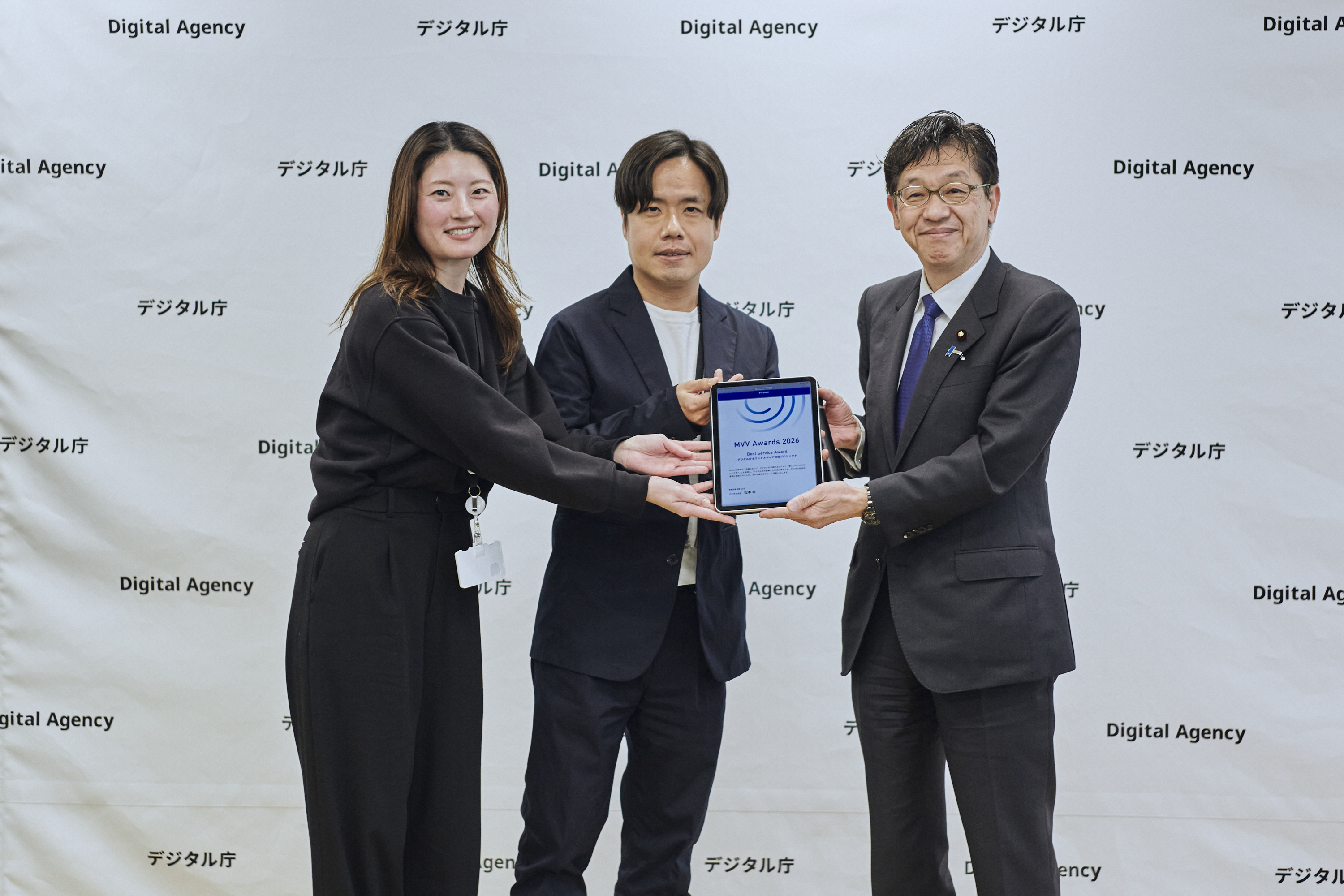 デジタル庁のバックボードを背景に、3名が「MVV Awards 2026 Best Service Award」の表彰状が表示されたタブレットを掲げて記念撮影している写真。