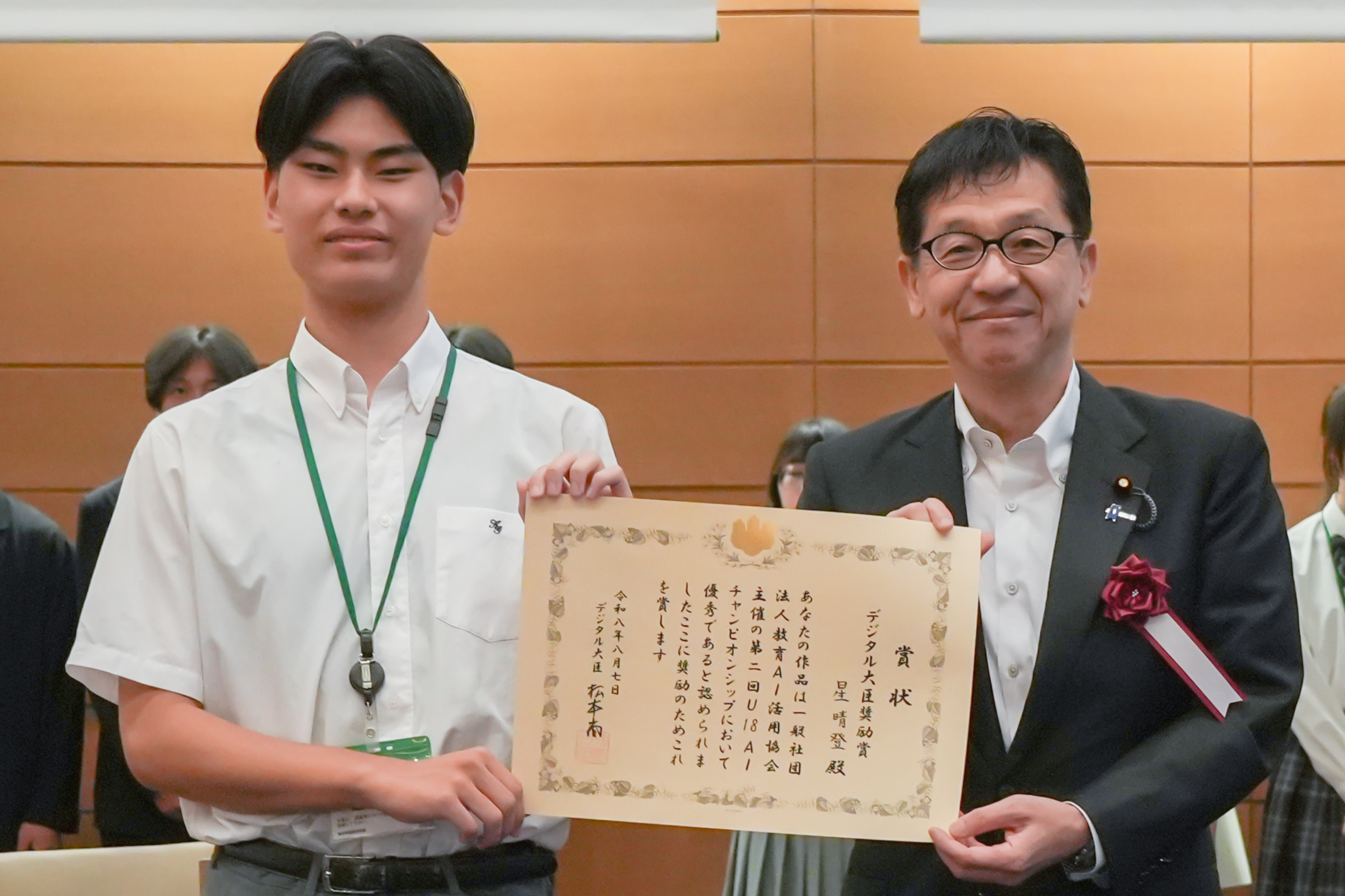 式典会場で、松本デジタル大臣と星さんが賞状を一緒に手にしながら正面に向かって微笑んでいる。左側の星さんは、白い半袖シャツに緑色のストラップのIDカードを首から下げている。右側の松本デジタル大臣は、ダークスーツに眼鏡をかけていて、胸に議員バッジと赤いコサージュを着用している。