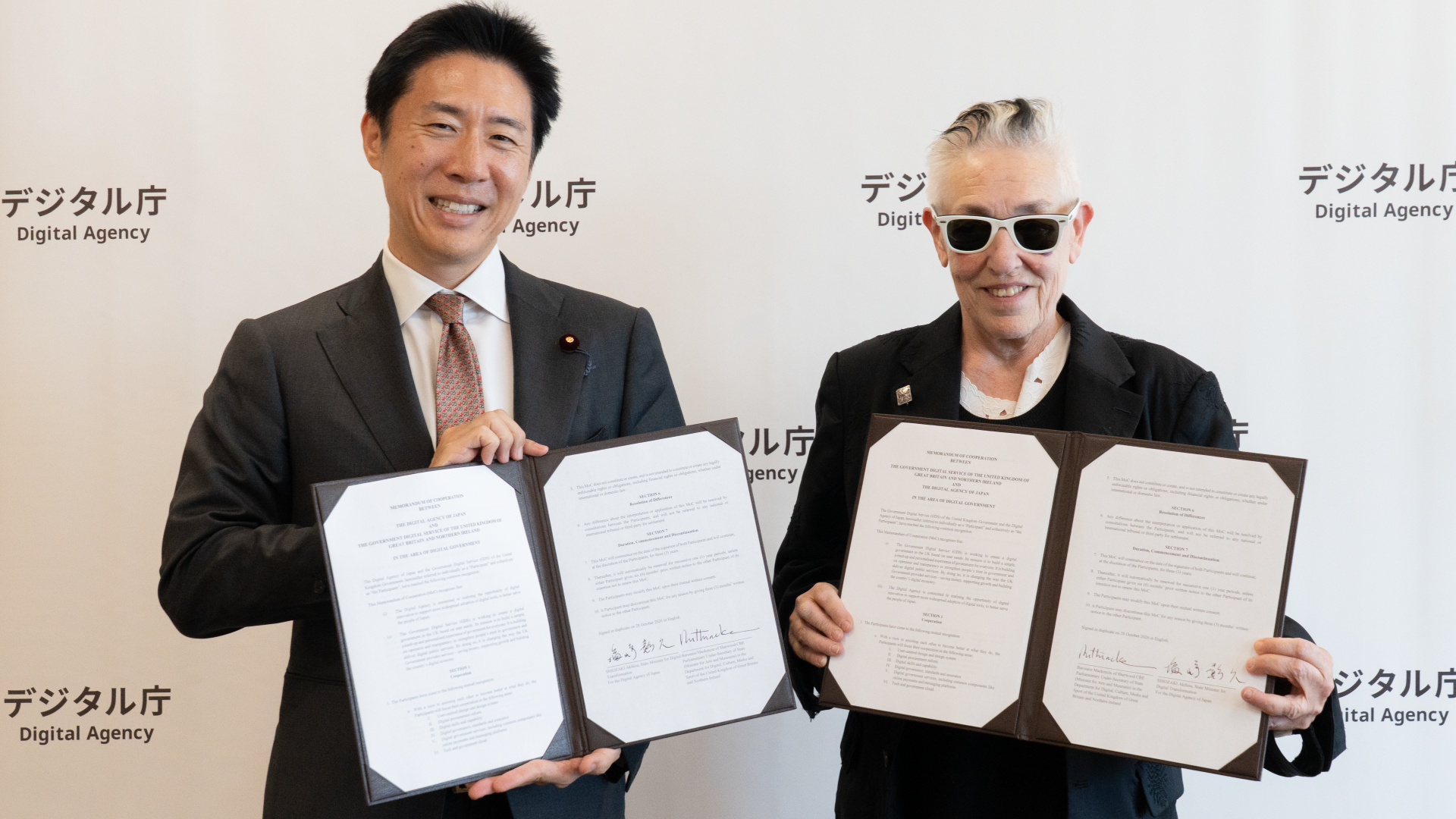 塩崎デジタル副大臣とマッケンジーDCMS閣外大臣が、それぞれ署名した文書を持ち笑顔で並んでいる。