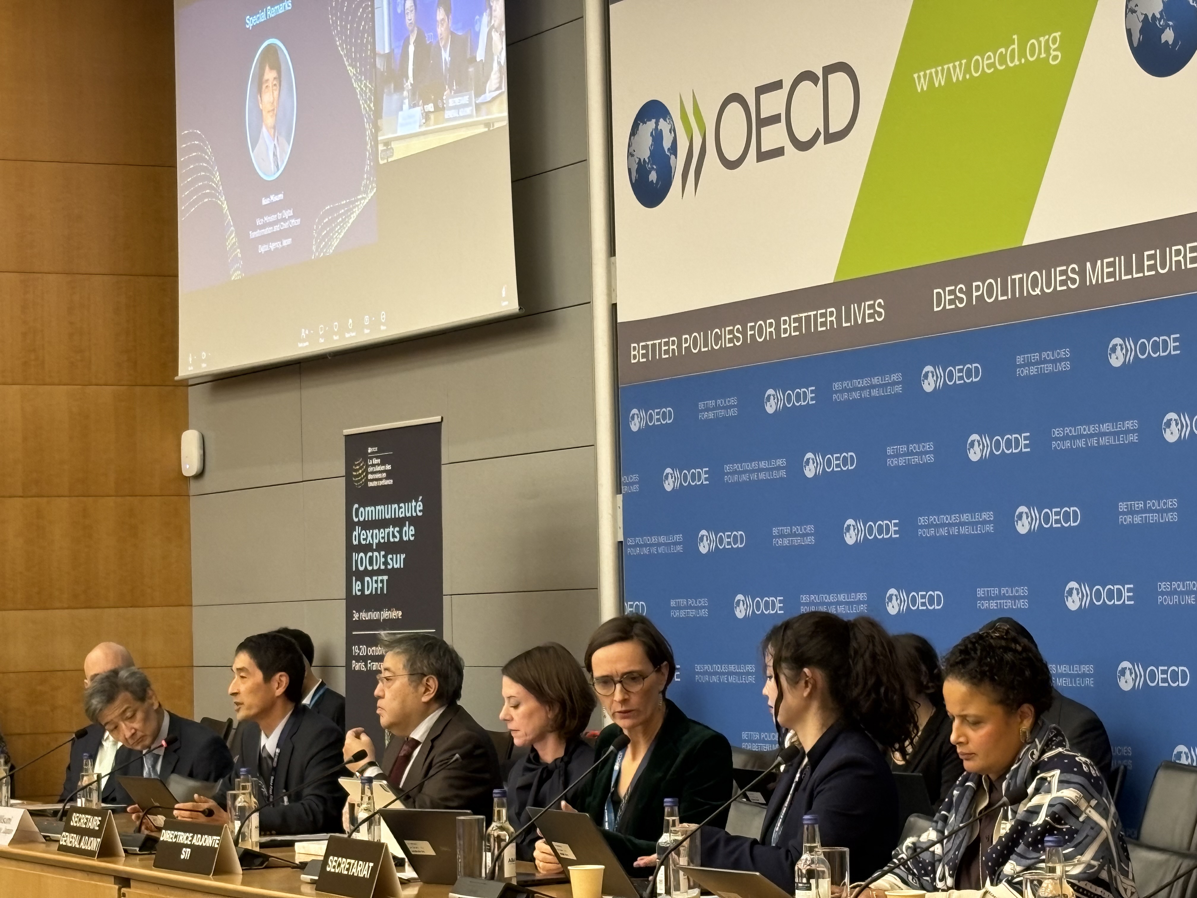 OECDのパネルの前の長机に出席者が7名ほどが並んで着席している。三角デジタル監は左から3番目で発言しており、上部のスクリーンにはその様子が映っている。