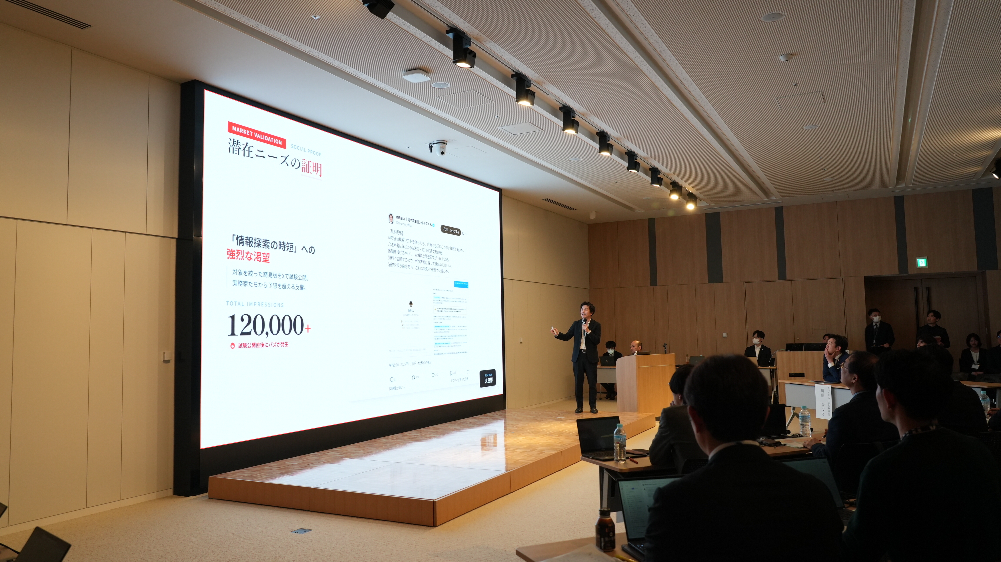 A hackathon participant gives a presentation on stage. A huge screen at the back of the stage shows introductory slides of the work. 