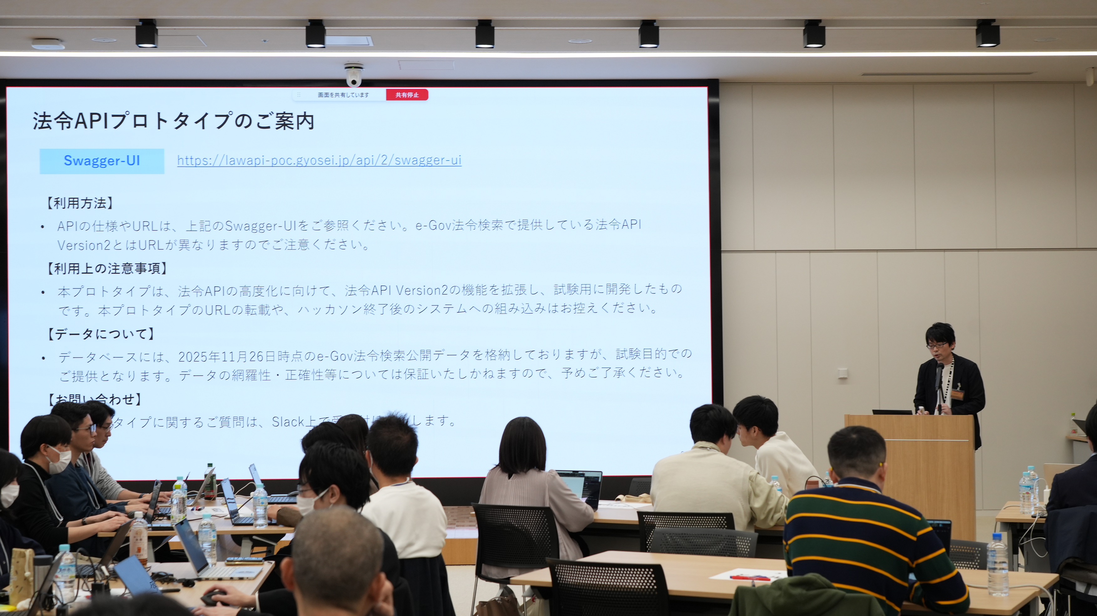 There is a large monitor in the front, and a Digital Agency official stands next to it and explains laws and regulations APIs to the participants. 