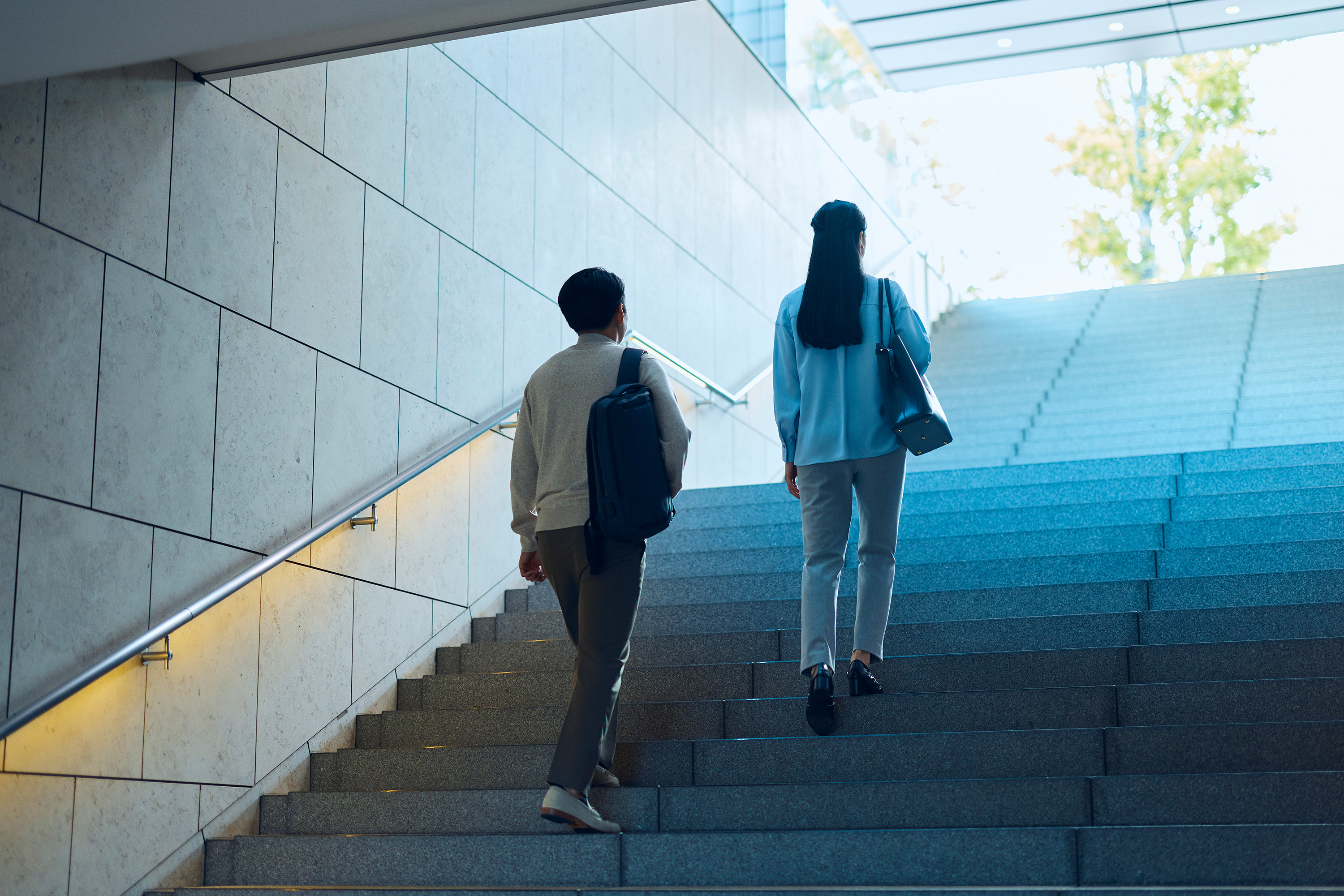 自然光が差し込むモダンな建物内の広い階段を上る2名。1名は水色のジャケットにハンドバッグ、もう1名はベージュのジャケットにリュックサック。