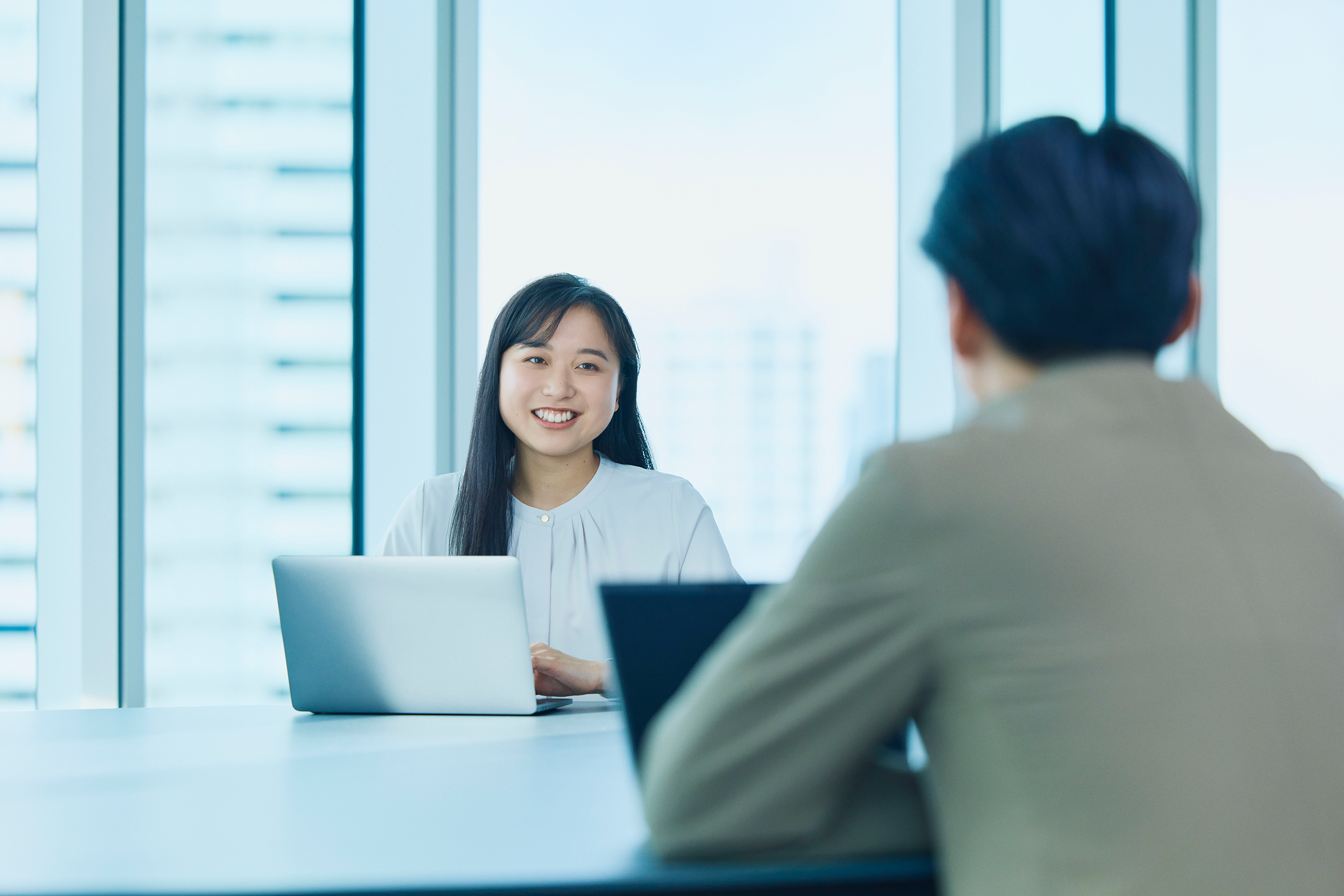 大きな窓のあるモダンなオフィスで、テーブルを挟んで向かい合いノートパソコンを使用する2名。面談またはインタビューの様子。