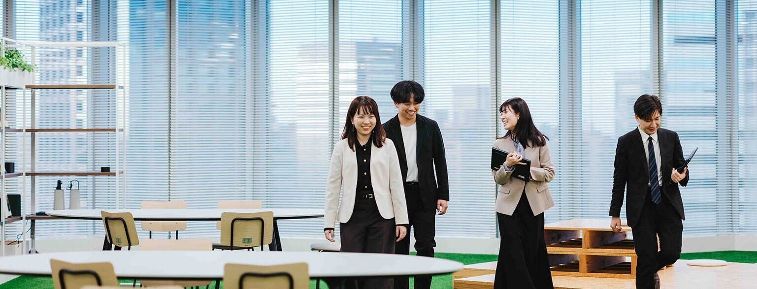 オフィスの広いスペースで4人のデジタル庁職員が歩いている写真。背景には大きな窓から都市の景色が見え、周辺には丸いテーブルと椅子が配置されている。床は緑色のカーペットで、天井には複数の照明がある