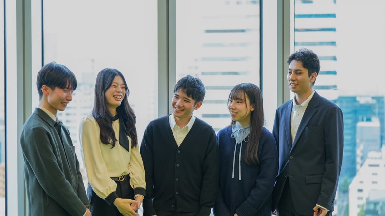 5人の人が写った写真。左側から男性、女性、男性、女性、男性と並んでおり、笑顔で談笑している。