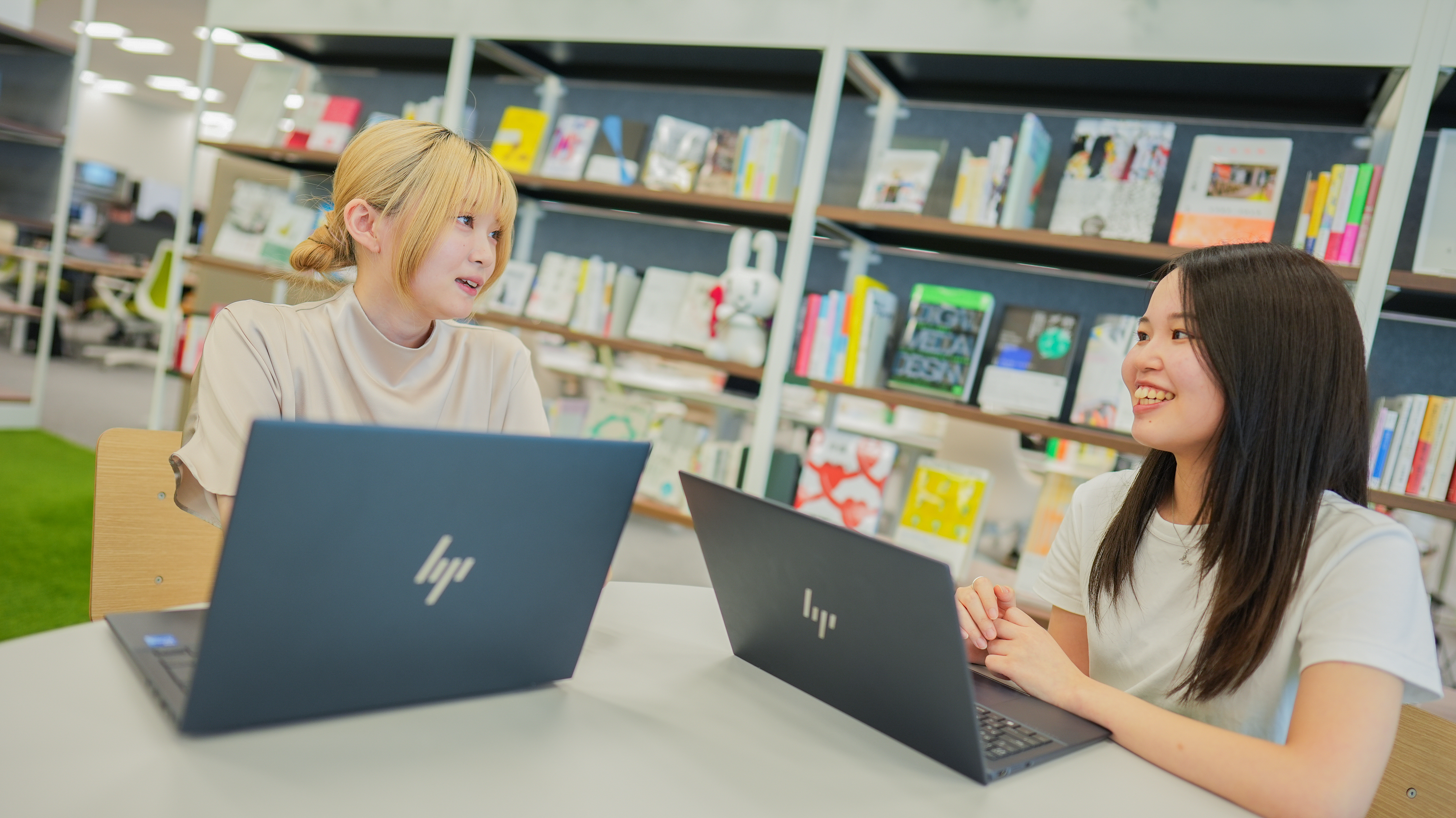 ノートパソコンを前に2人が会話している写真。背景には本棚があり、本が並んでいる。