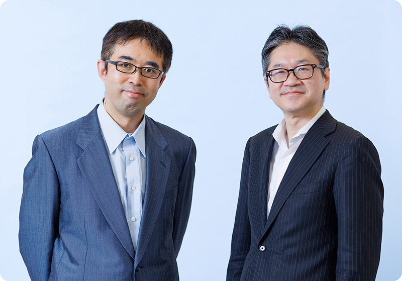 大塚参事官と上田参事官が並んで写っているバストショット。二人ともスーツを着てノーネクタイである。