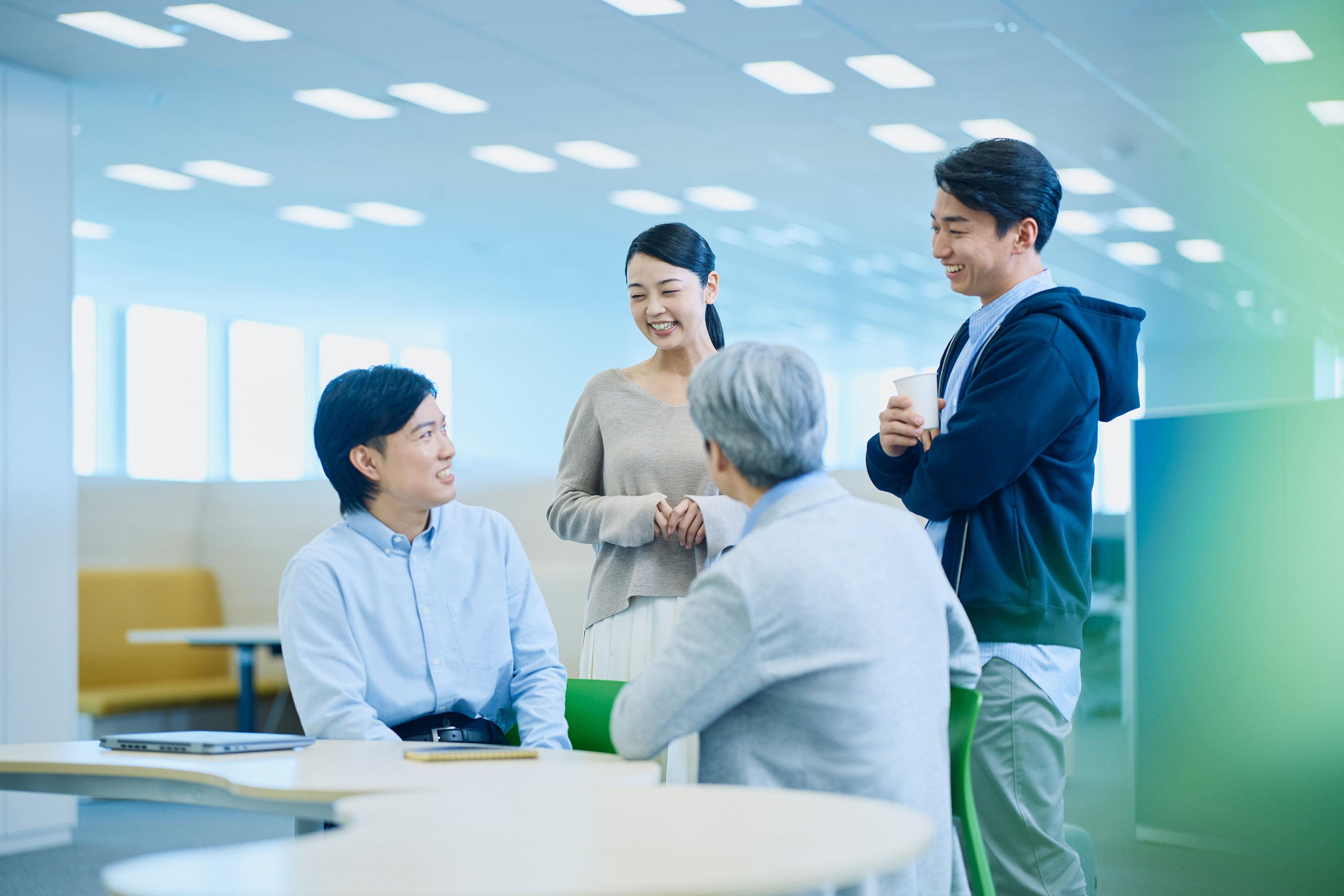 明るく開放的なオフィス空間で、4人のビジネスパーソンが立ったまま和やかに会話している写真。左側には水色のシャツを着た若い男性が座っており、中央にはベージュのニットを着た女性が笑顔で立っている。右側には水色のシャツと紺色のパーカーを着た男性がカップを持って立っており、手前にはグレーのジャケットを着た白髪の男性が後ろ姿で写っている。背景には青と緑を基調とした現代的なオフィスデザインで、天井には白い照明が規則的に配置され、黄色いソファが見える。