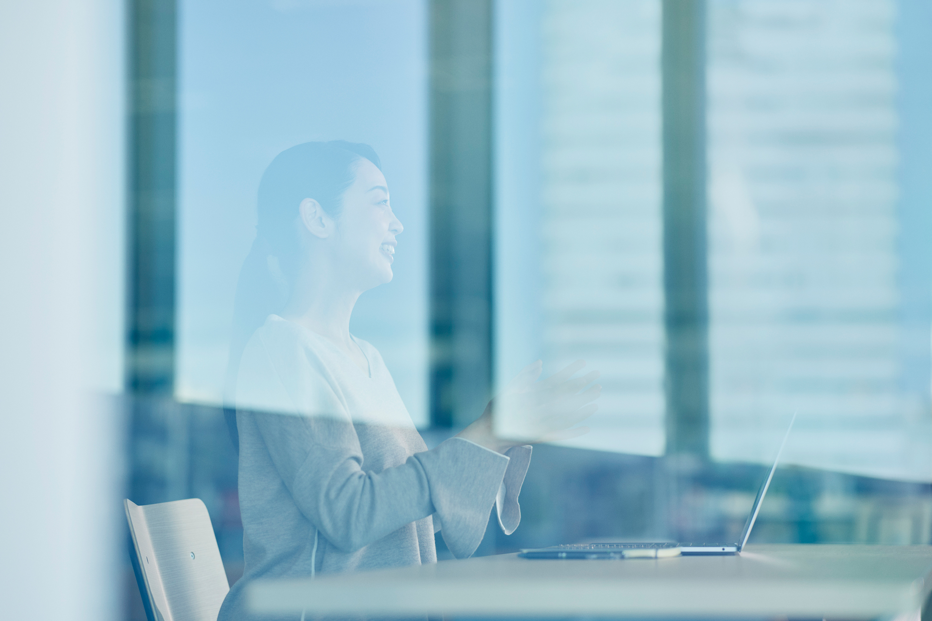 オフィス内の一角、ガラス越しにベージュのニットを着た女性が笑顔で話している写真。デスクの上にはノートパソコンが置かれており、ガラスには高層ビルが映り込んでいる。全体に青みがかった明るい光が差し込んでいる。