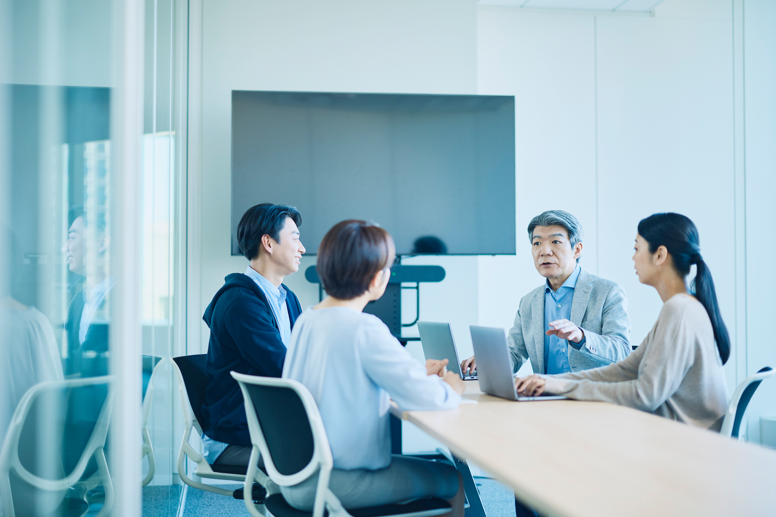 ガラスの壁のある明るく開放的な会議室で、4人のビジネスパーソンが木製のテーブルを囲んで議論している写真。左側には水色のシャツと紺色のパーカーを着た若い男性がにこやかな表情で座り、中央手前には水色のブラウスを着た女性が背中を向けて座っている。右側にはグレーのジャケットを着た中年男性がノートパソコンを前に、身振りを交えながら説明している様子。その隣ではベージュのニットを着た女性がノートパソコンを操作している。背景には大型のモニターが設置されている。