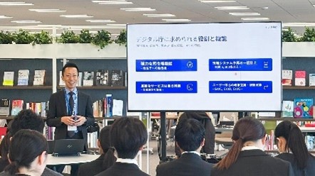 業務説明会の様子の写真。前方に大きなモニターがあり、その隣に人が立っており、聴講者に向かって笑顔で話している。