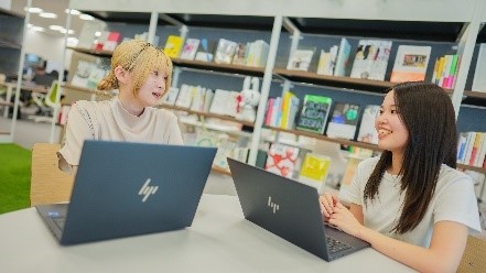 ノートパソコンを前に2人が会話している写真。背景には本棚があり、本が並んでいる。