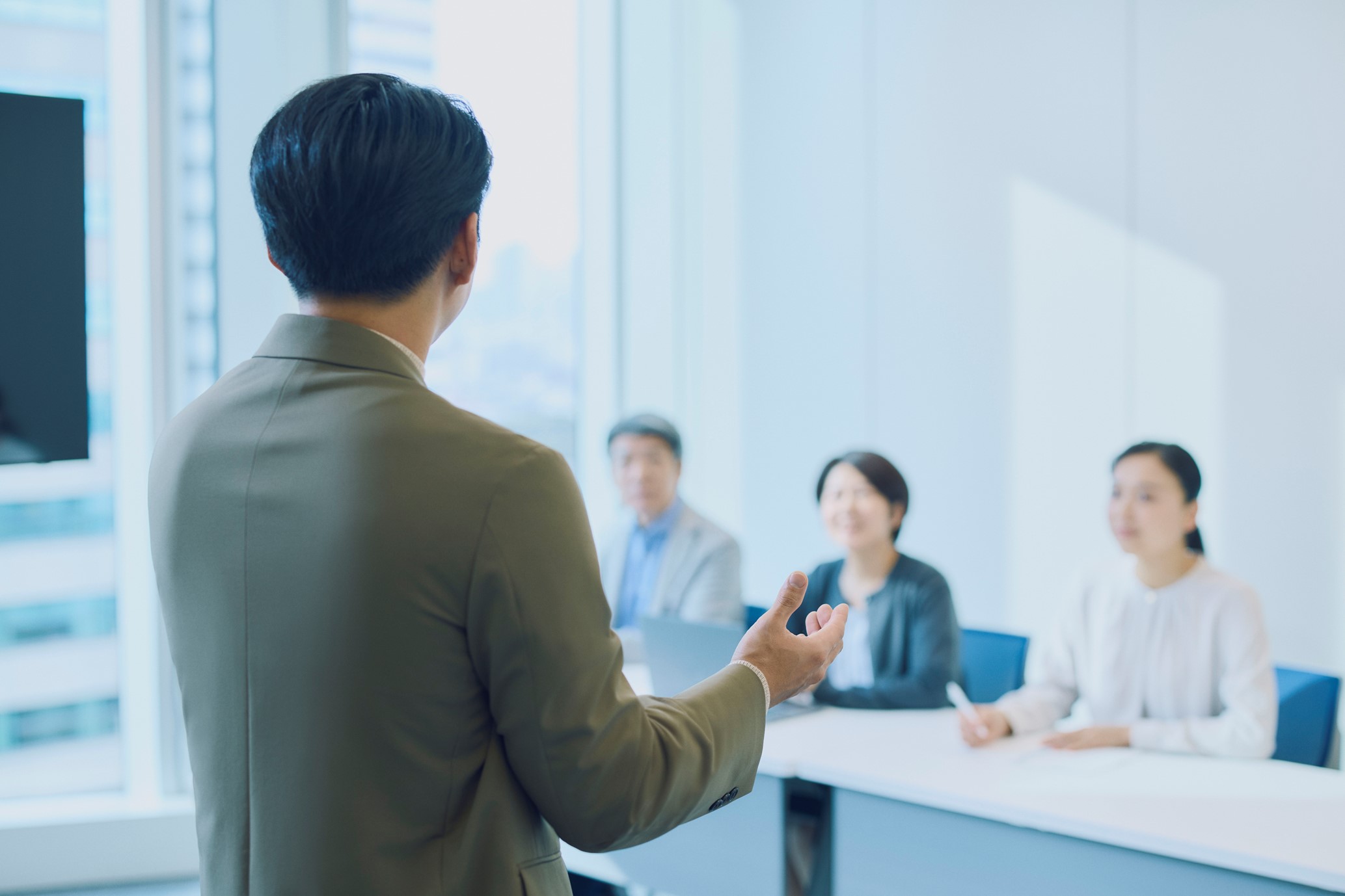 スーツを着た人物が着席した3名に向けてプレゼンテーションを行っているビジネス会議の写真。発表者は片手でジェスチャーをしながら説明しており、聴衆は真剣に耳を傾けている。公式な議論またはブリーフィングの場であることが伺える。