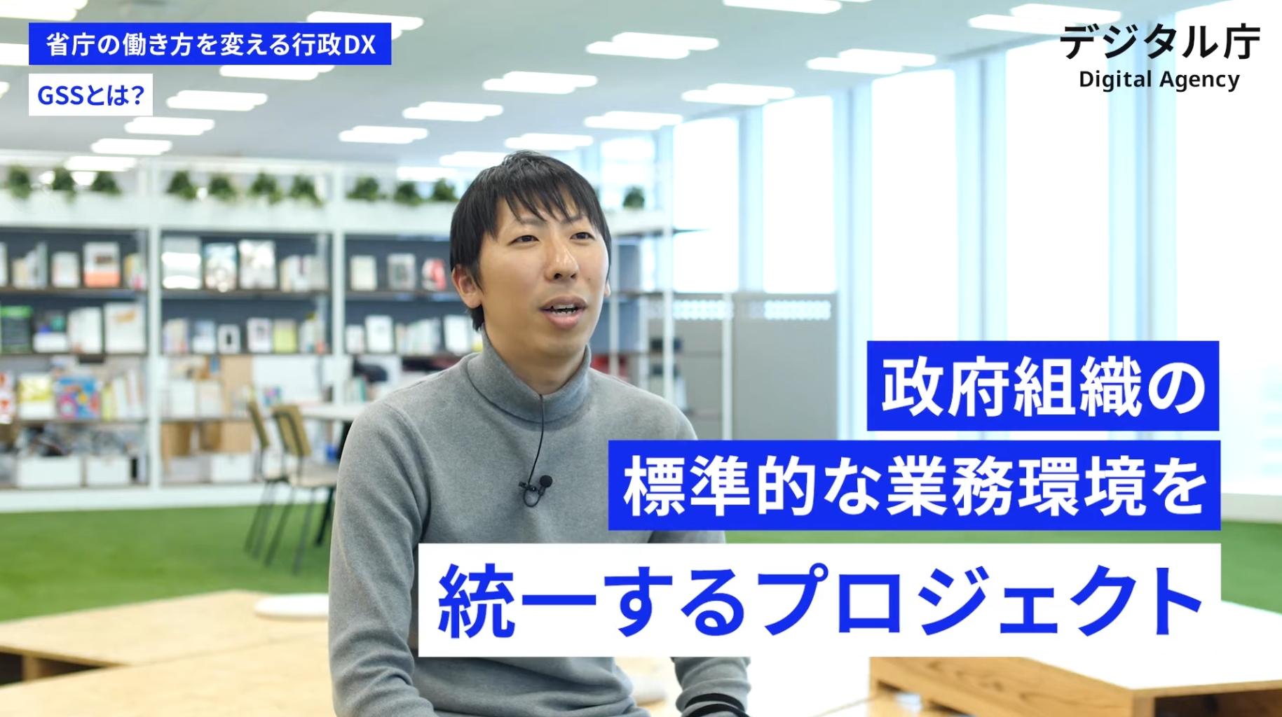 デジタル庁のロゴと日本語テキストが掲載された写真。本棚と植物のあるモダンなオフィスに座る人物が写っており、政府組織内の業務環境の標準化・統一化に関するプロジェクトを紹介している。