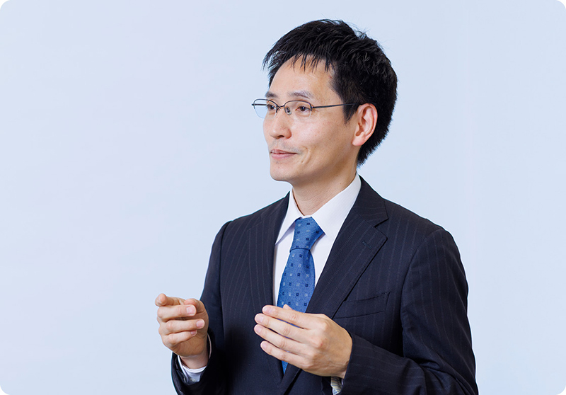 加藤企画官のバストショット。紺色のスーツを着て青いネクタイを締めている。軽く両手を上げて誰かに話そうとしている。