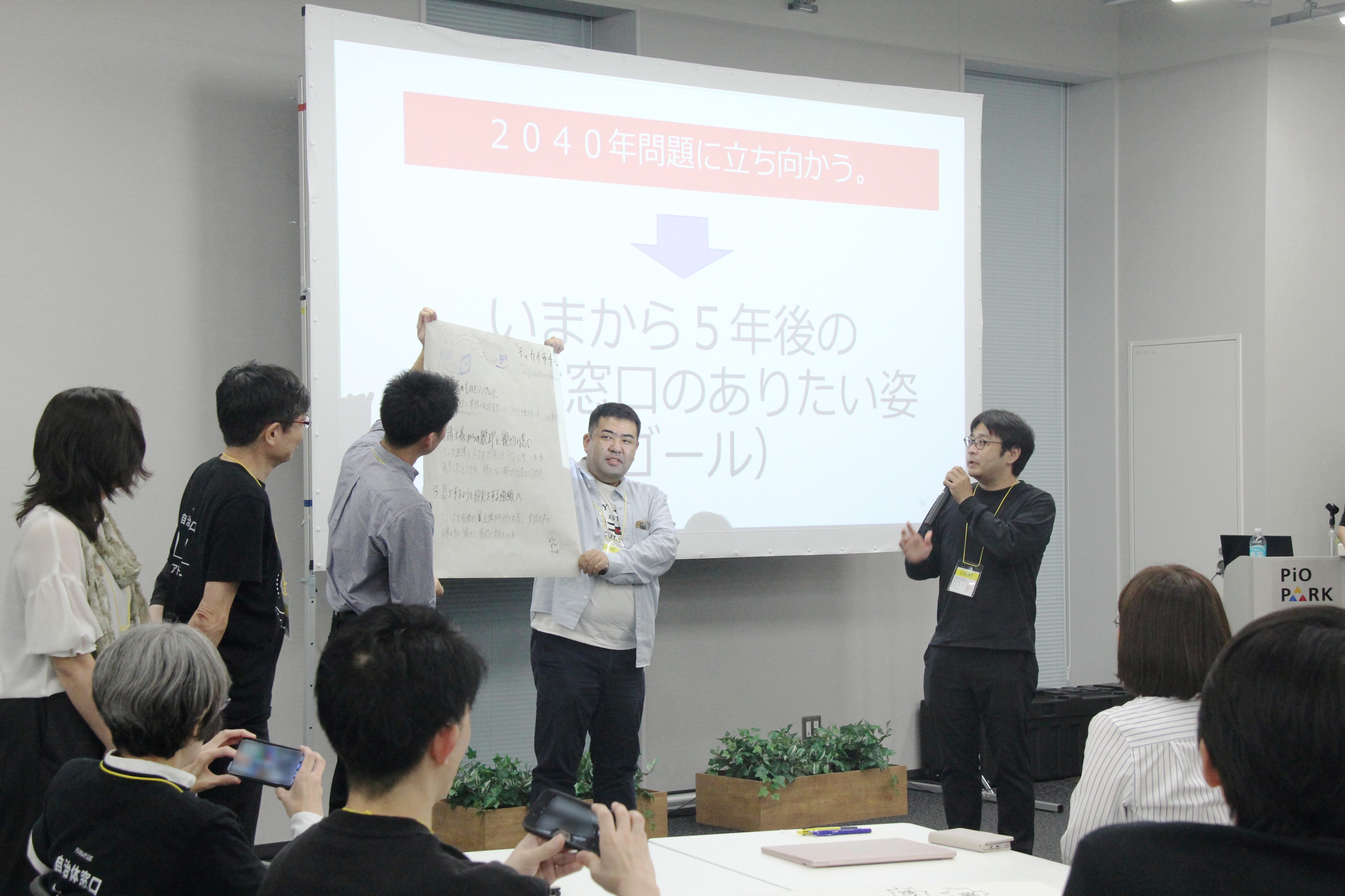 5人のグループがスクリーンの前に横に並んで立っている。うち2人は発表内容をまとめた模造紙を掲げており、右手の男性はマイクを手に説明をしている。