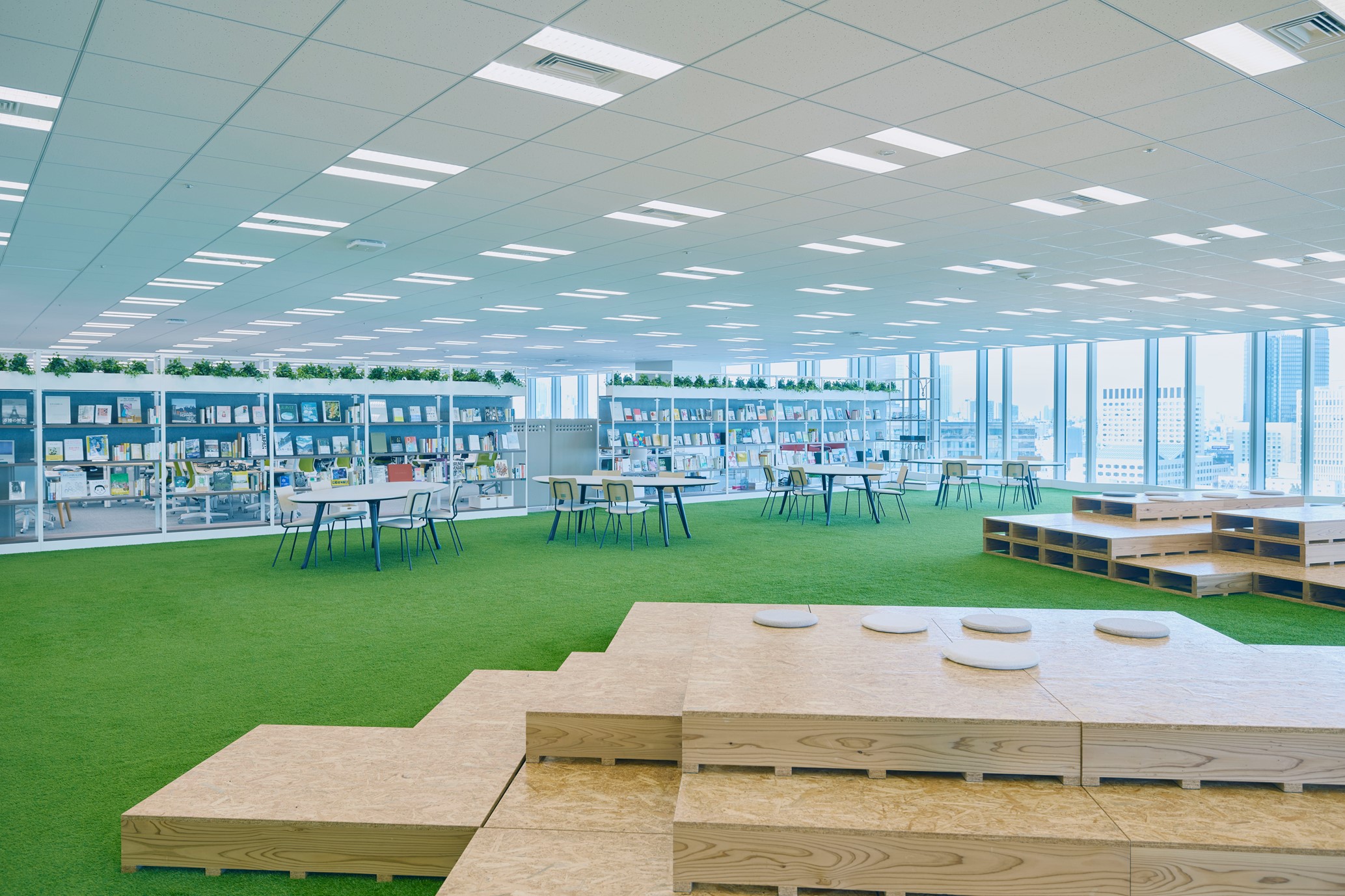 大きな窓、緑のカーペットの床、丸いクッションが置かれた木製の段状座席を備えた広々としたデジタル庁芝生スペースの写真。背面の壁には本が並んだ棚が設置されており、テーブルと椅子が点在している。明るく開放的な環境が印象的なスペースとなっている。