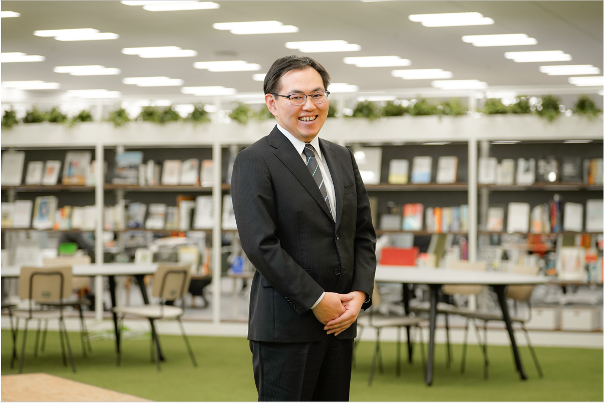 A male official is pictured standing in his office, with bookshelves, tables and chairs arranged in an orderly fashion in the background. 