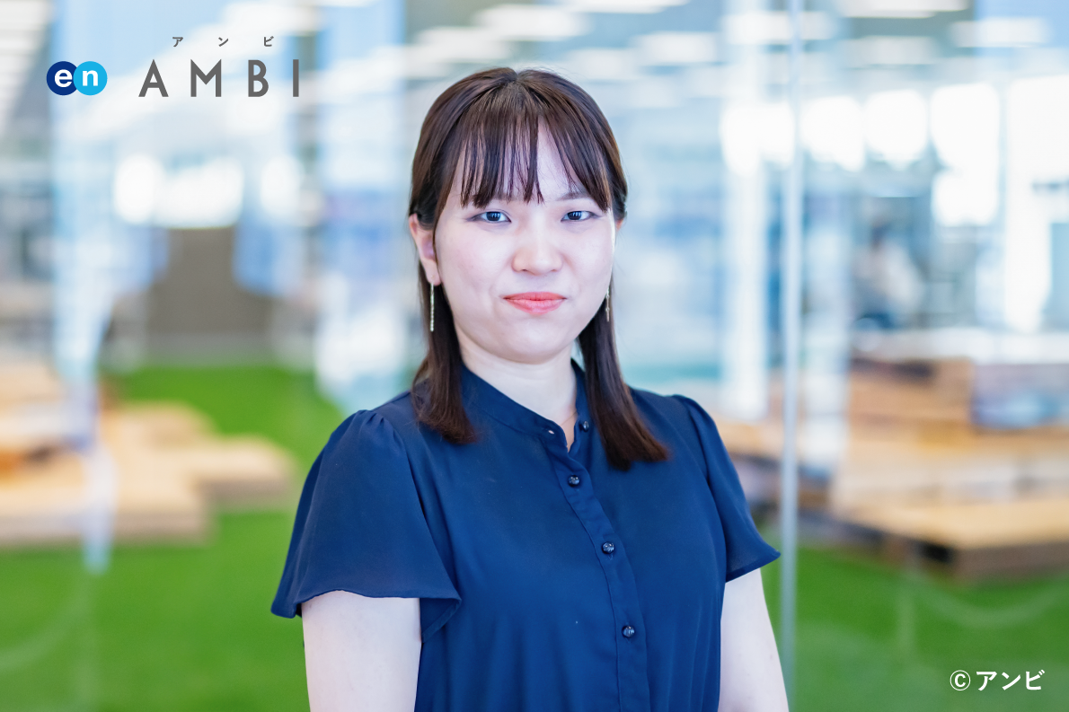 A female official is pictured standing inside an indoor office, with the "en AMBI" logo in the upper left, indicating a workplace or office environment. 