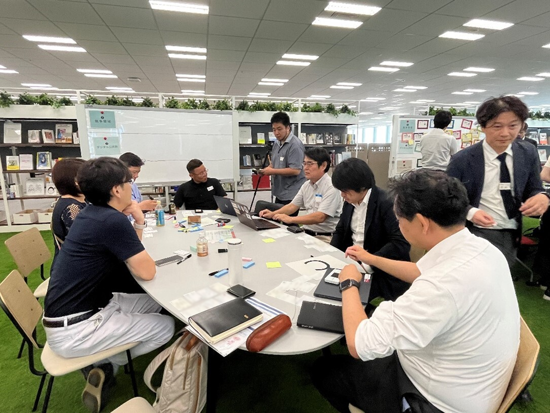 円卓に参加者6人が向かい合って座り、パソコンやふせんなどを使いながらディスカッションをしている写真
