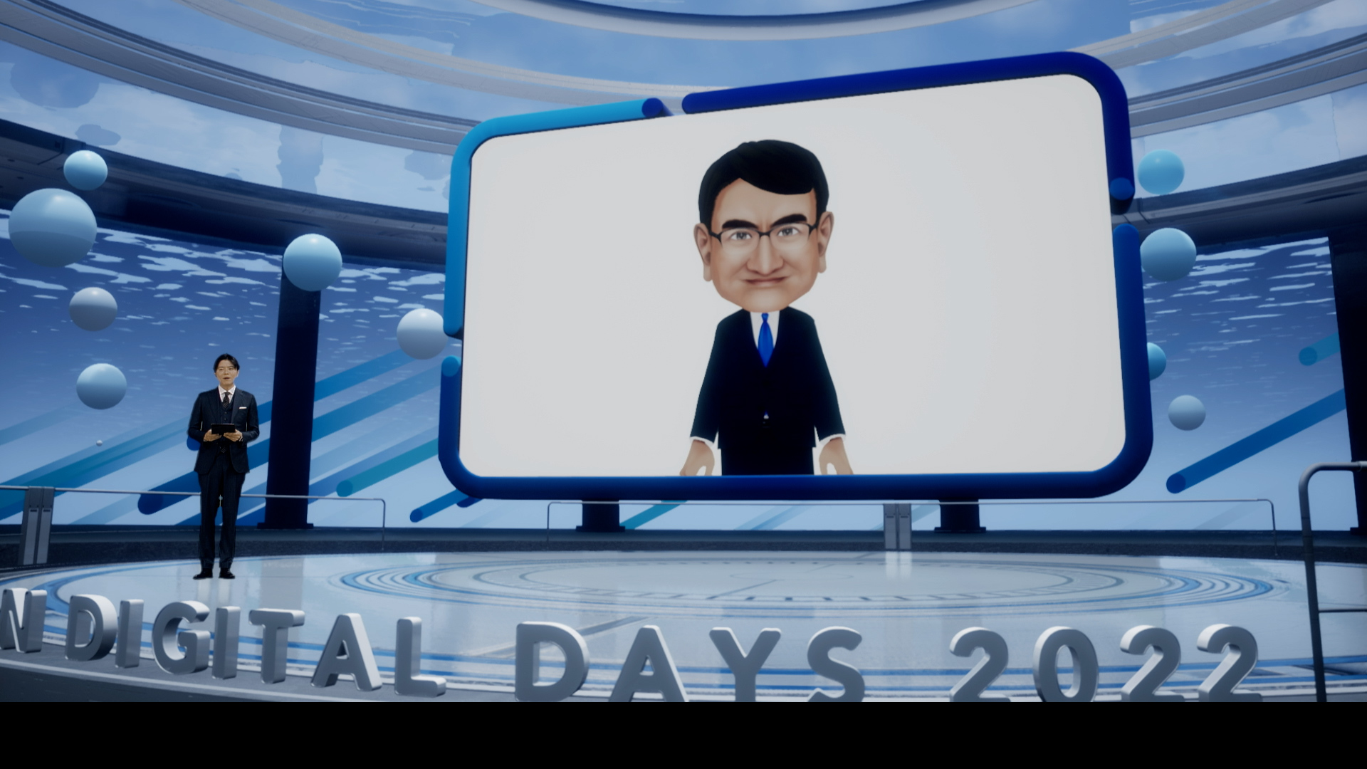 Digital Day An on-line event venue with an announcer standing on the left side of the virtual studio and a large monitor in the center of the stage showing a Minister Kono digital avatar.