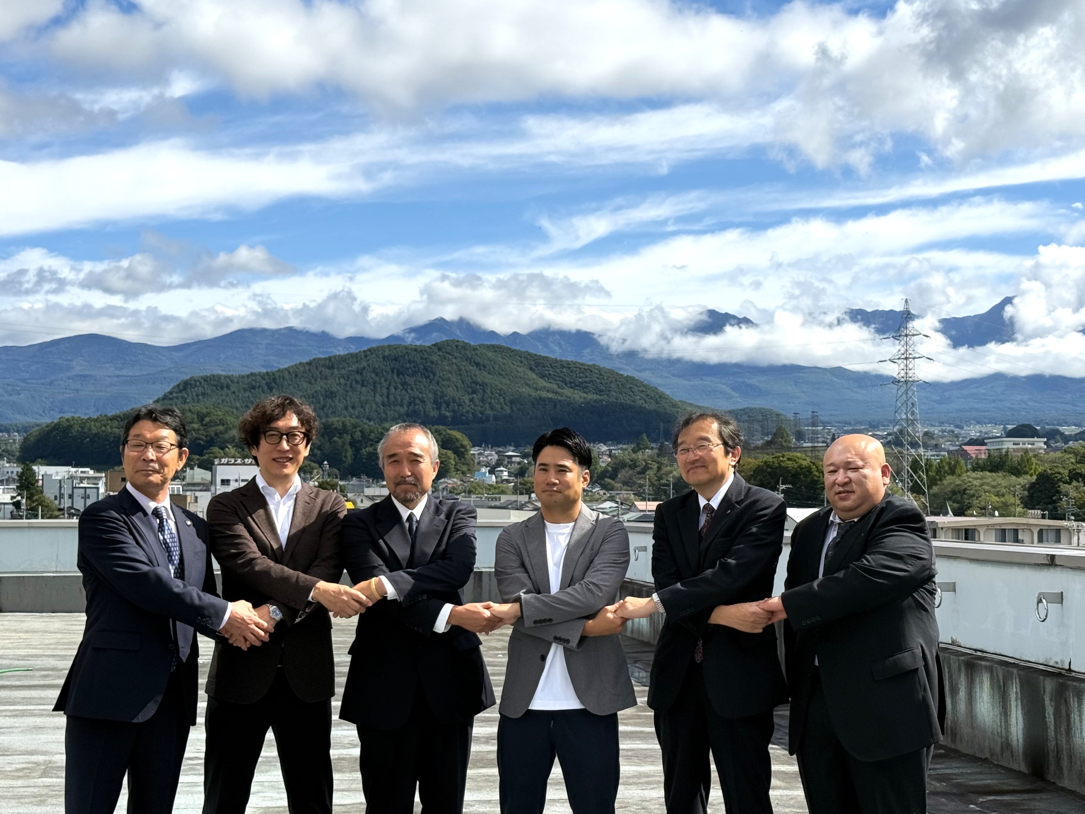 長野県茅野市役所の屋外にて、山々と青空を背景に左から茅野市 今井敦市長、デジタル庁国民向けサービスグループ鳥山企画調整官、八ヶ岳観光協会 兼 黒百合ヒュッテ 米川岳樹代表、北アルプス山小屋友交会 兼 槍ヶ岳山荘グループ 穂苅大輔代表、茅野市 熊谷晃地域創生政策監、八ヶ岳観光協会副会長 兼 唐沢鉱泉 両角岩男代表が並び、腕を交差しながら両隣同士握手をしている