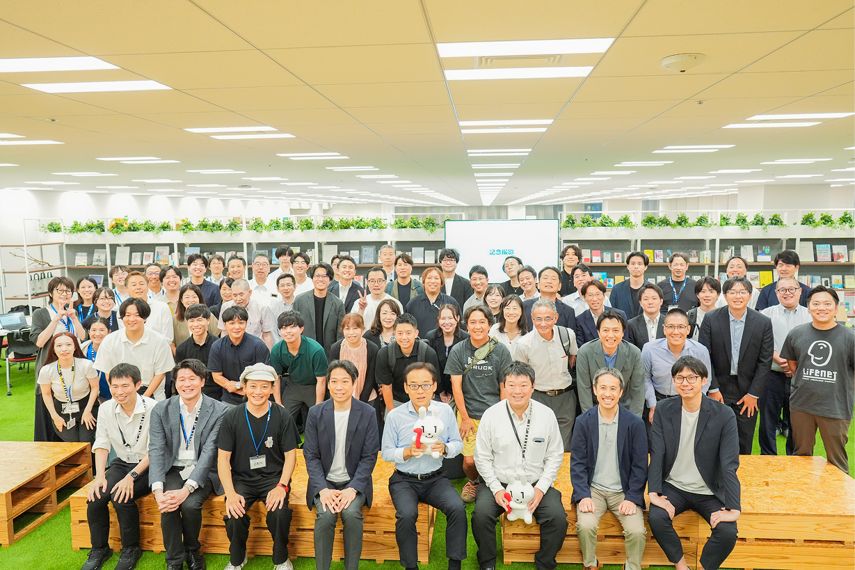 書棚と緑の人工芝が配置されたオフィスのスペースにて、約60名が5列ほど並んだ集合写真