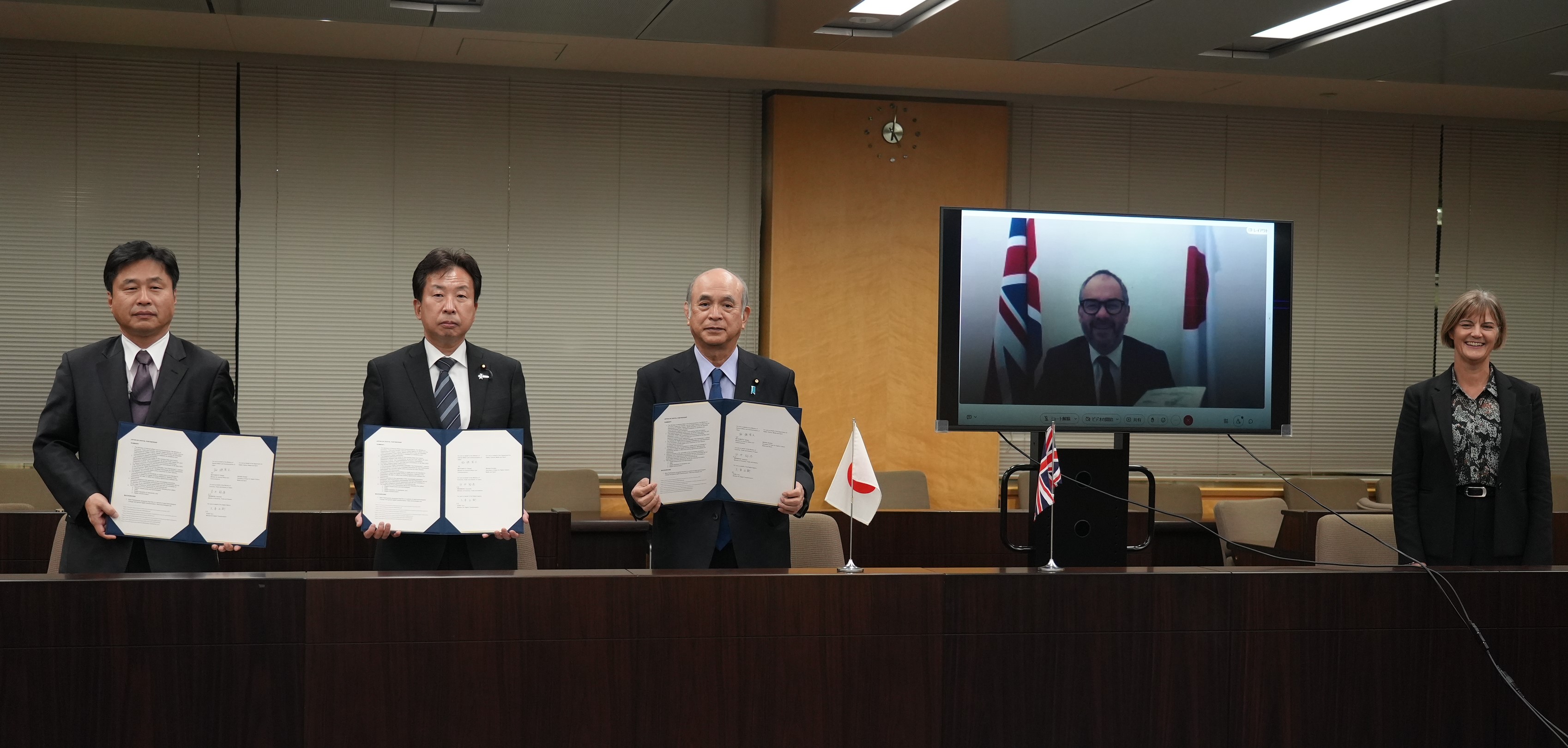 Photo of 4 people. From left to right, Mr. Hirai, Vice Minister for International Affairs at the Ministry of Economy, Trade and Industry, Mr. Ogushi, State Minister for Digital Transformation, Mr. Tsuge, State Minister for Internal Affairs and Communications; Mr. Scully, Parliamentary Under Secretary of State at the Department for Digital, Culture, Media and Sport shown in the screen. Everyone in the photo is holding the agreements in their hands.