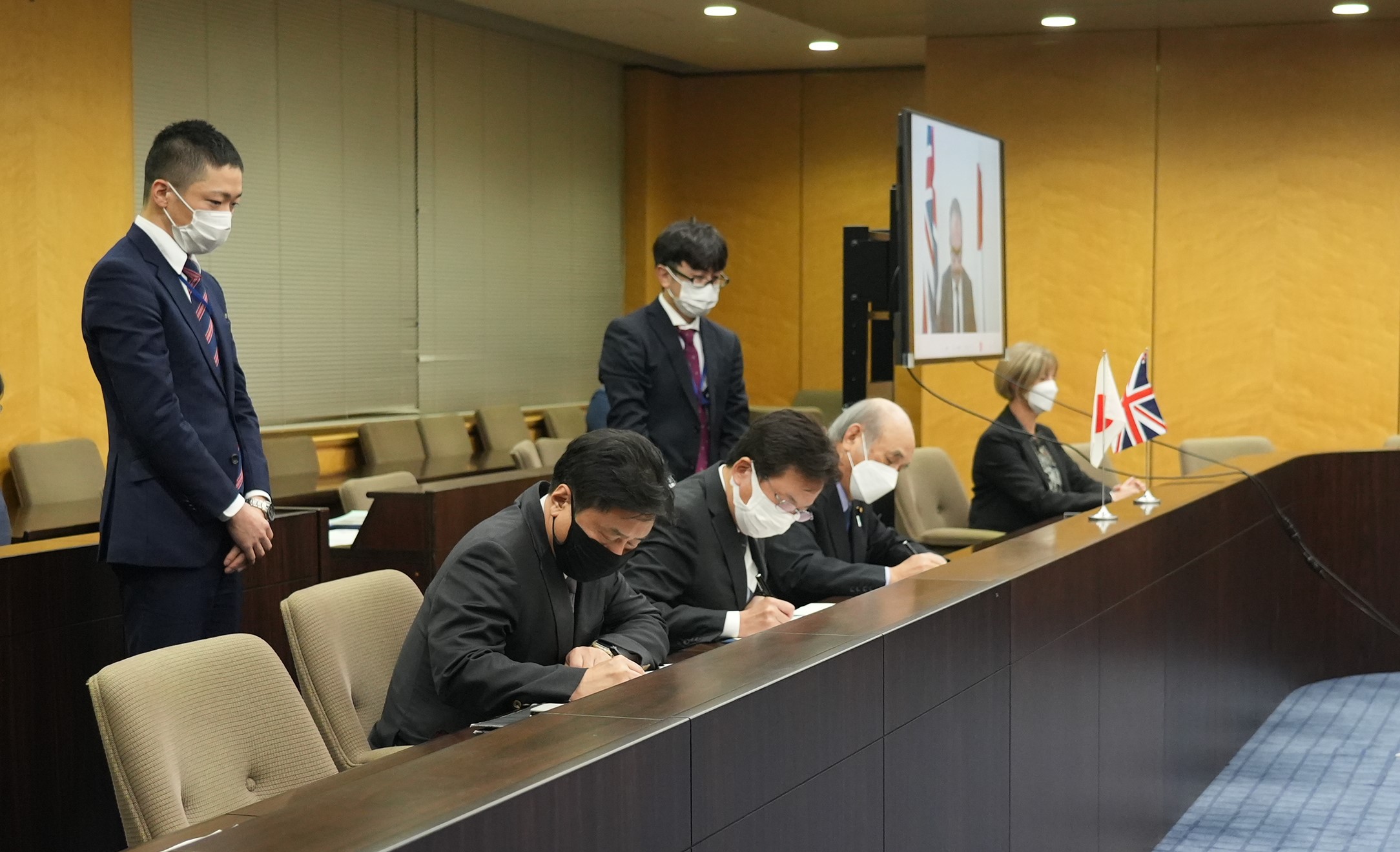 左から、平井経済産業審議官、大串デジタル副大臣、柘植総務副大臣、テレビに映されたスカリーDCMS政務次官が並んで机に向かって座っている。4名とも合意文書にサインをしている。