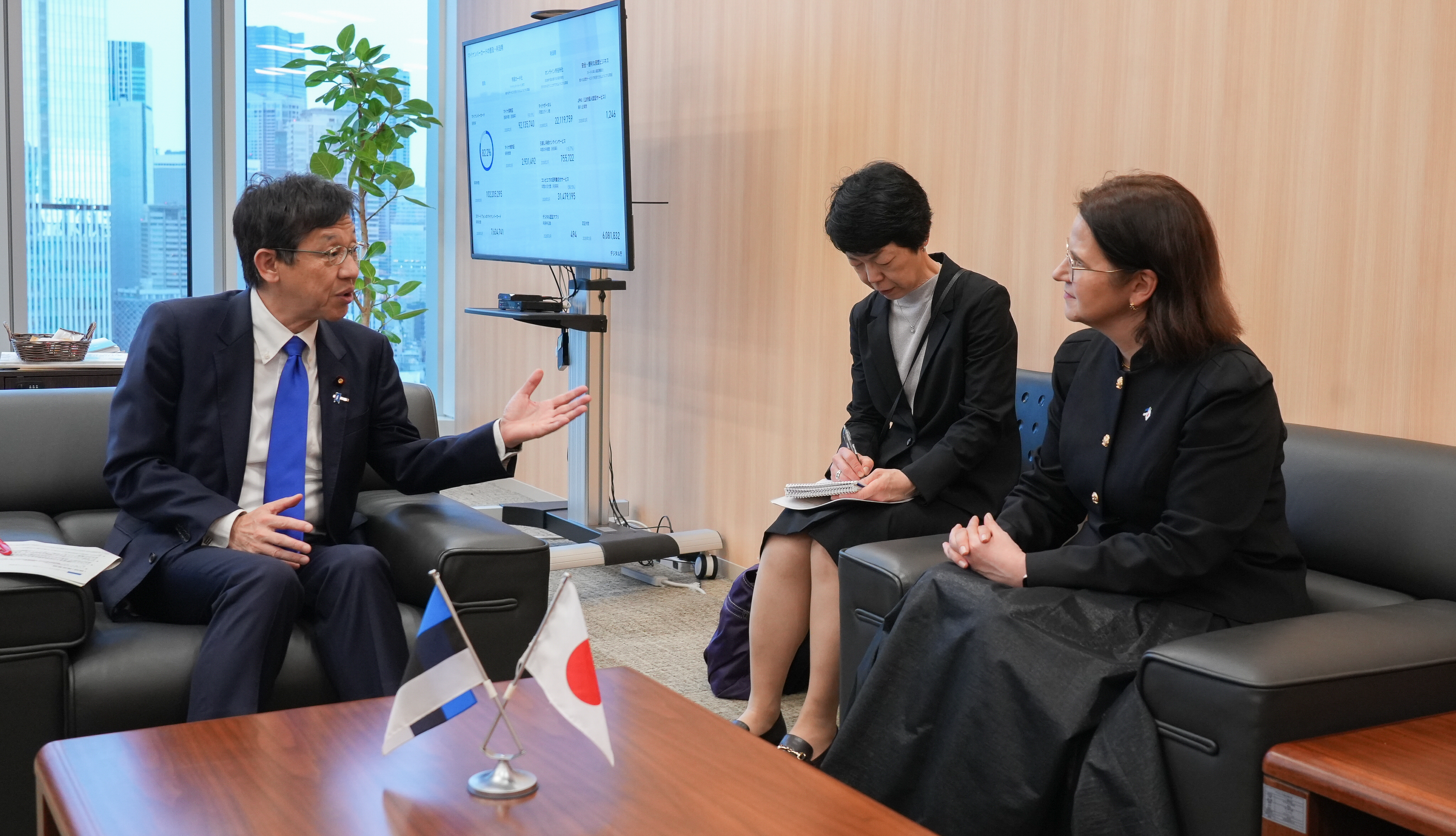 室内の応接スペースで、日本とエストニアの小型国旗が置かれたテーブルを挟んで、3名が会談している。左側には濃紺のスーツに青いネクタイを着用した松本デジタル大臣が身振り手振りを交えながら話しており、右側には黒いジャケット姿のリーサ＝リー・パコスタ法務・デジタル大臣が向かい合って座っている。中央には黒い服装の女性がノートを手にメモを取っており、通訳または記録担当と見られる。背後には大型モニターが設置され、図表が映し出されている。窓の外には高層ビル群の景色が広がっている。