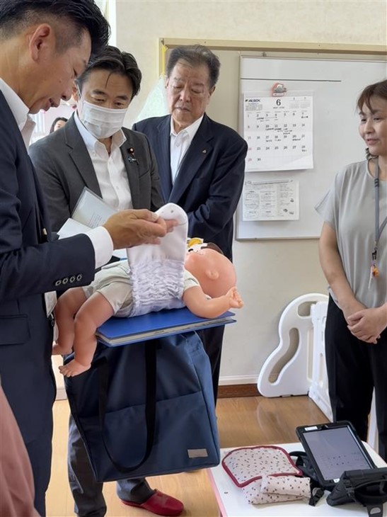幼児の人形に着衣型のIoTを装着している様子を見る穂坂副大臣の写真。