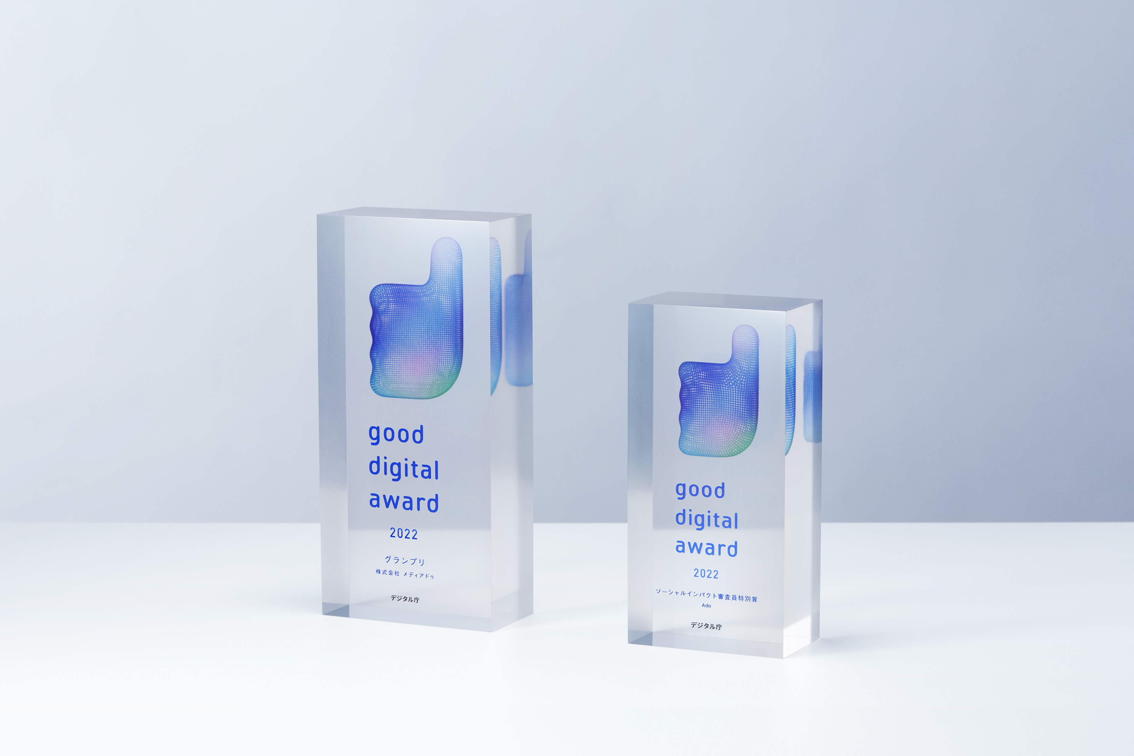 good digital awardのトロフィー2つの3DCGイメージ。トロフィーは透明な直方体の中心からやや上の箇所にgood digital awardのロゴマークである親指を抽象化したマークが埋め込まれている。左側の直方体の表面にはgood digital award 2022グランプリ株式会社メディアドゥデジタル庁というテキストが7行になった状態で刻印されており、右側の直方体の表面にはgood digital award 2022ソーシャルインパクト審査員特別賞Adoデジタル庁というテキストが単語ごとに改行されて7行になった状態で刻印されている。大きさの違う2つのトロフィーは横に並べられていて、正面よりやや斜め方向から見たアングルのCGになっている。