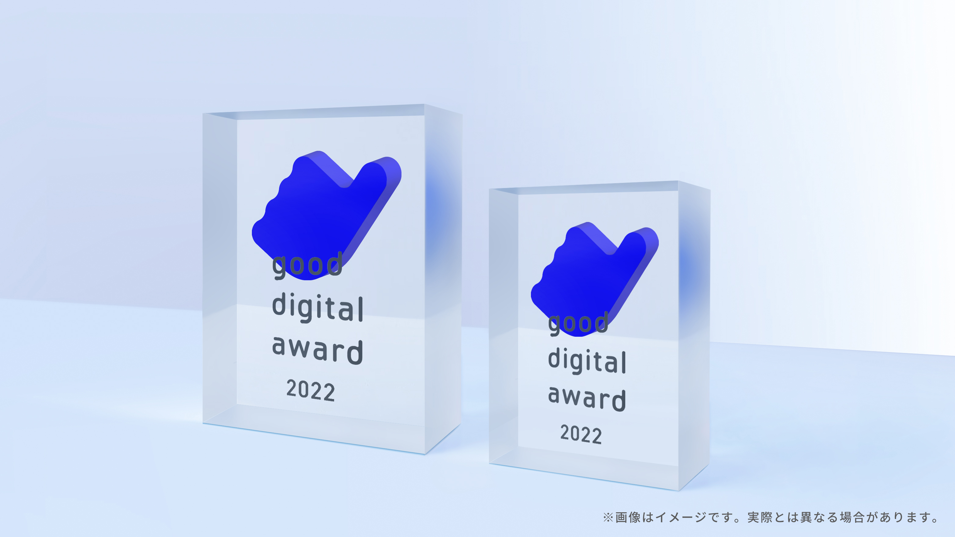 good digital awardのトロフィー2つの3DCGイメージ。トロフィーは透明な直方体の中心からやや上の箇所にgood digital awardのロゴマークである親指を抽象化したマークが埋め込まれている。直方体の表面にはgood digital award 2022というテキストが単語ごとに改行されて4行になった状態で刻印されている。大きさの違う2つのトロフィーは横に並べられていて、正面よりやや斜め方向から見たアングルのCGになっている。