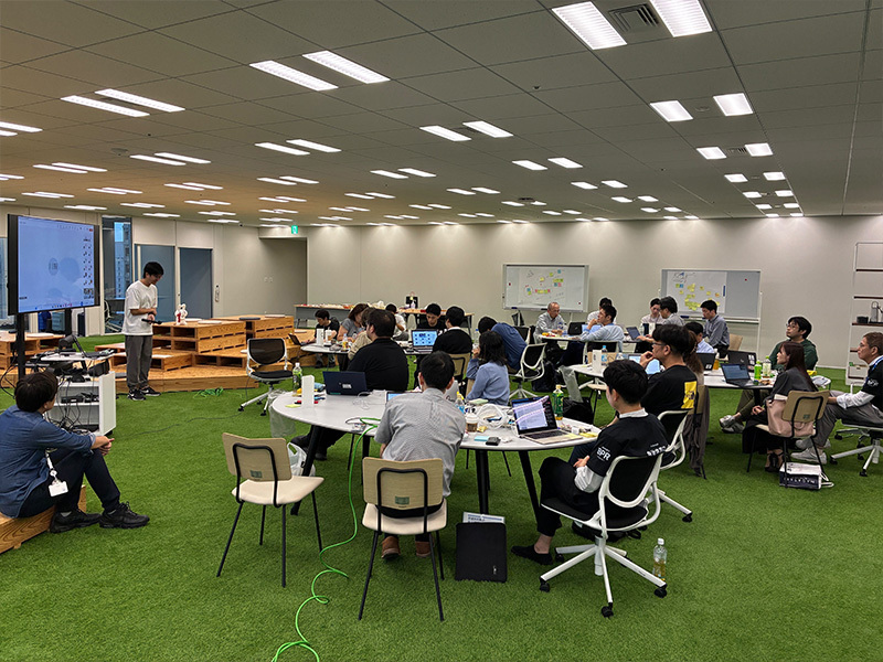 広い会場の前方で発表者がモニター横に立ち話している様子の写真。発表者の前には複数の丸いテーブルが並んでおり、参加者が座って聞いています。