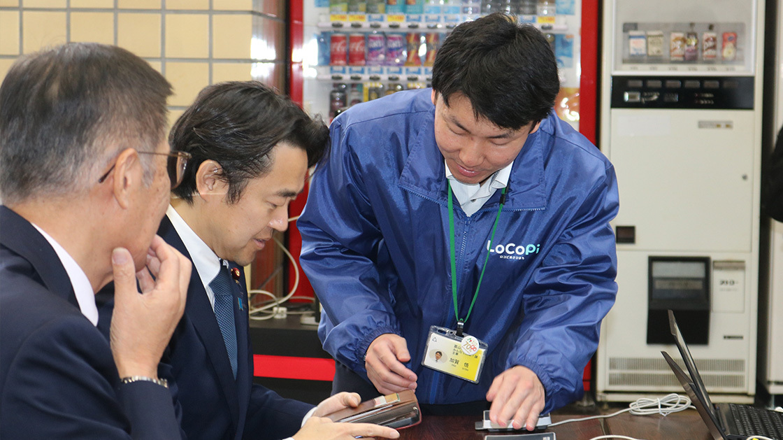 穂坂デジタル副大臣が朝日町職員の説明を受けながら、スマートフォンを使いLoCoPiあさひまち利用登録をしている様子。