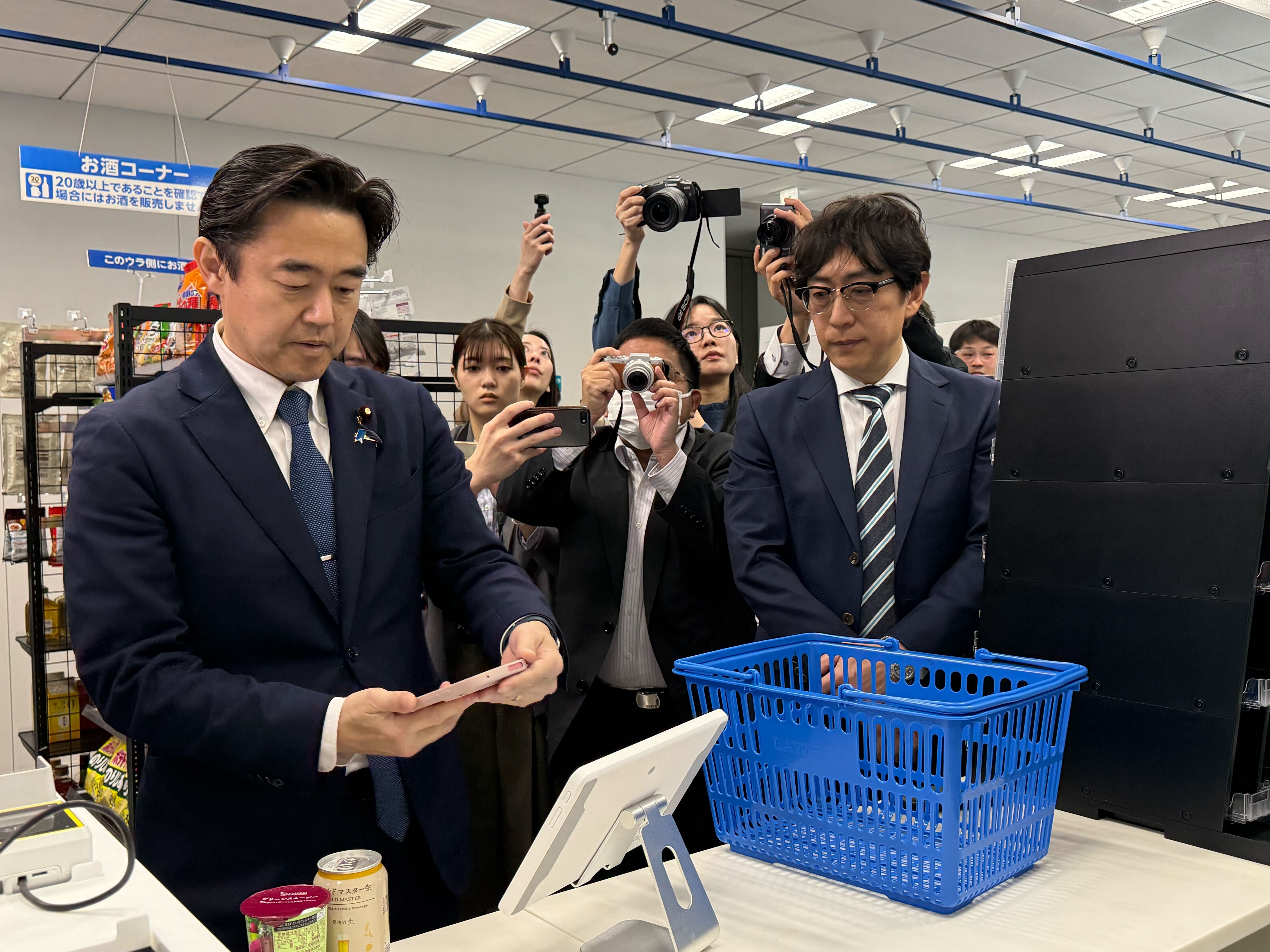 Mr. Hosaka, State Minister for Digital Transformation, on the left, a tablet with a self-checkout function in the center, and Mr. Hosaka, State Minister for Digital Transformation, holding a smartphone with a My Number Card function over the tablet
