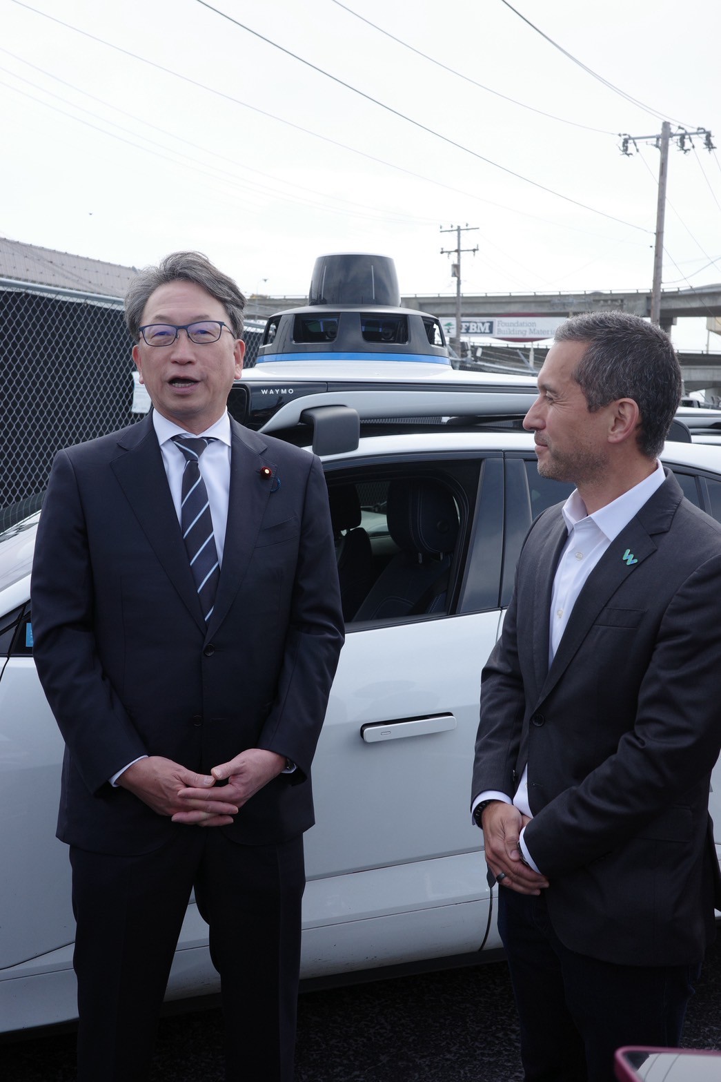自動運転車の前で、平デジタル大臣と自動運転タクシー企業担当者が立って話している様子