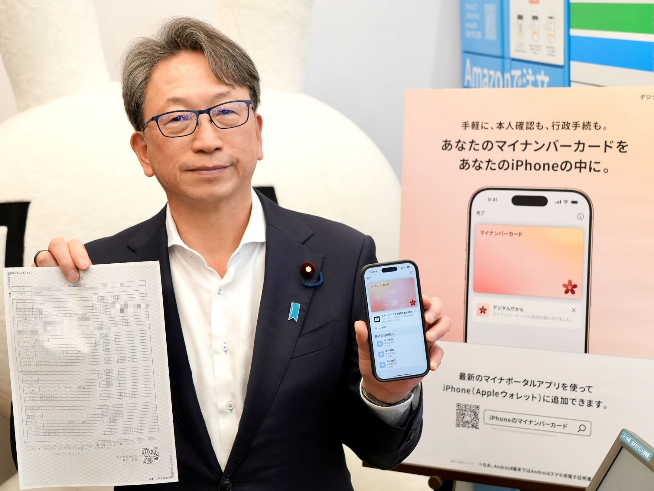A photo of Mr. Taira, Minister for Digital Transformation, holding a copy of his certificate of residence printed from a multi-copy machine in his right hand and an iPhone with My Number Card on it in his left hand