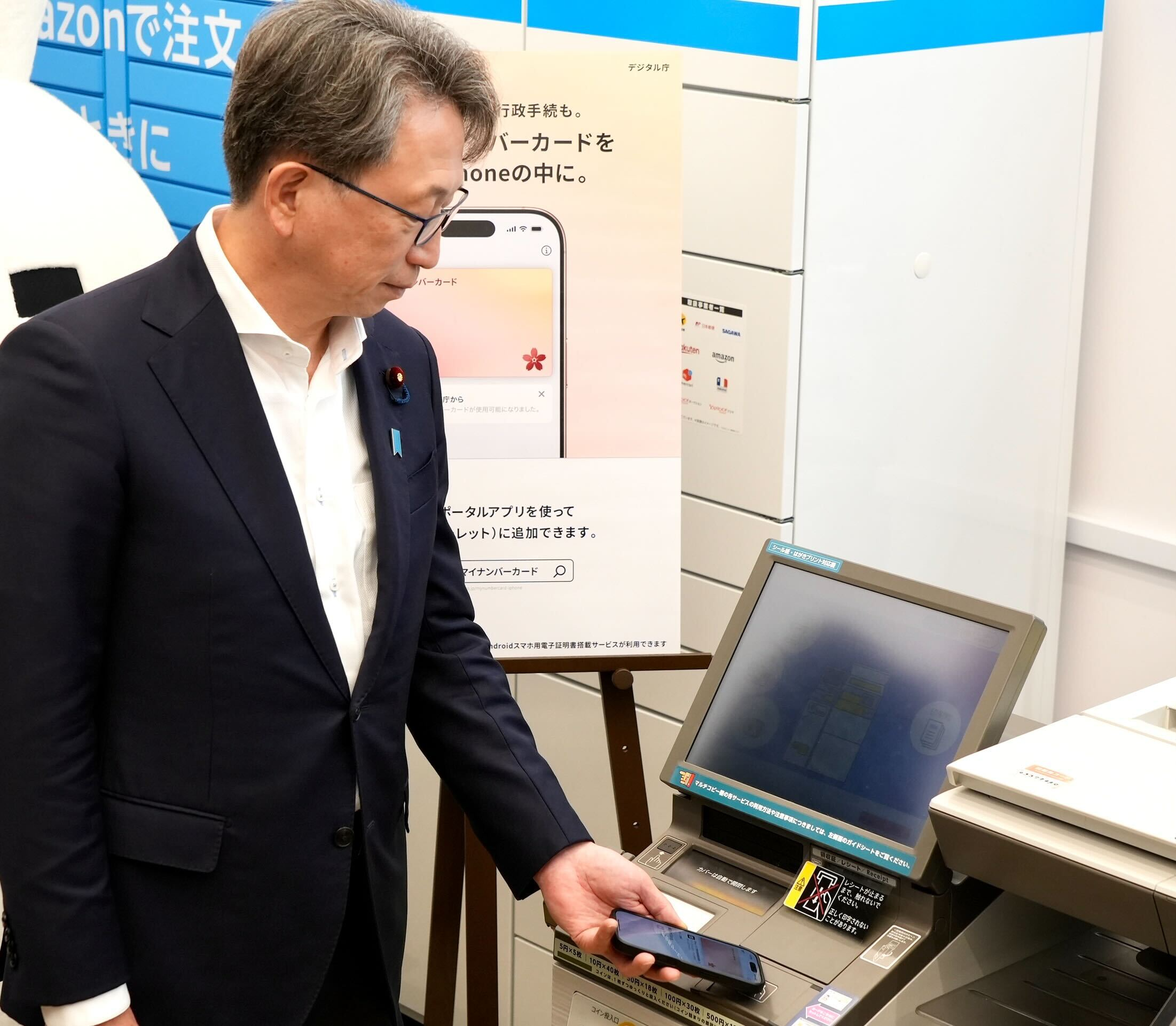 Photo of Mr. Taira, Minister for Digital Transformation, holding his phone in his left hand and holding it up to a multi-copy machine