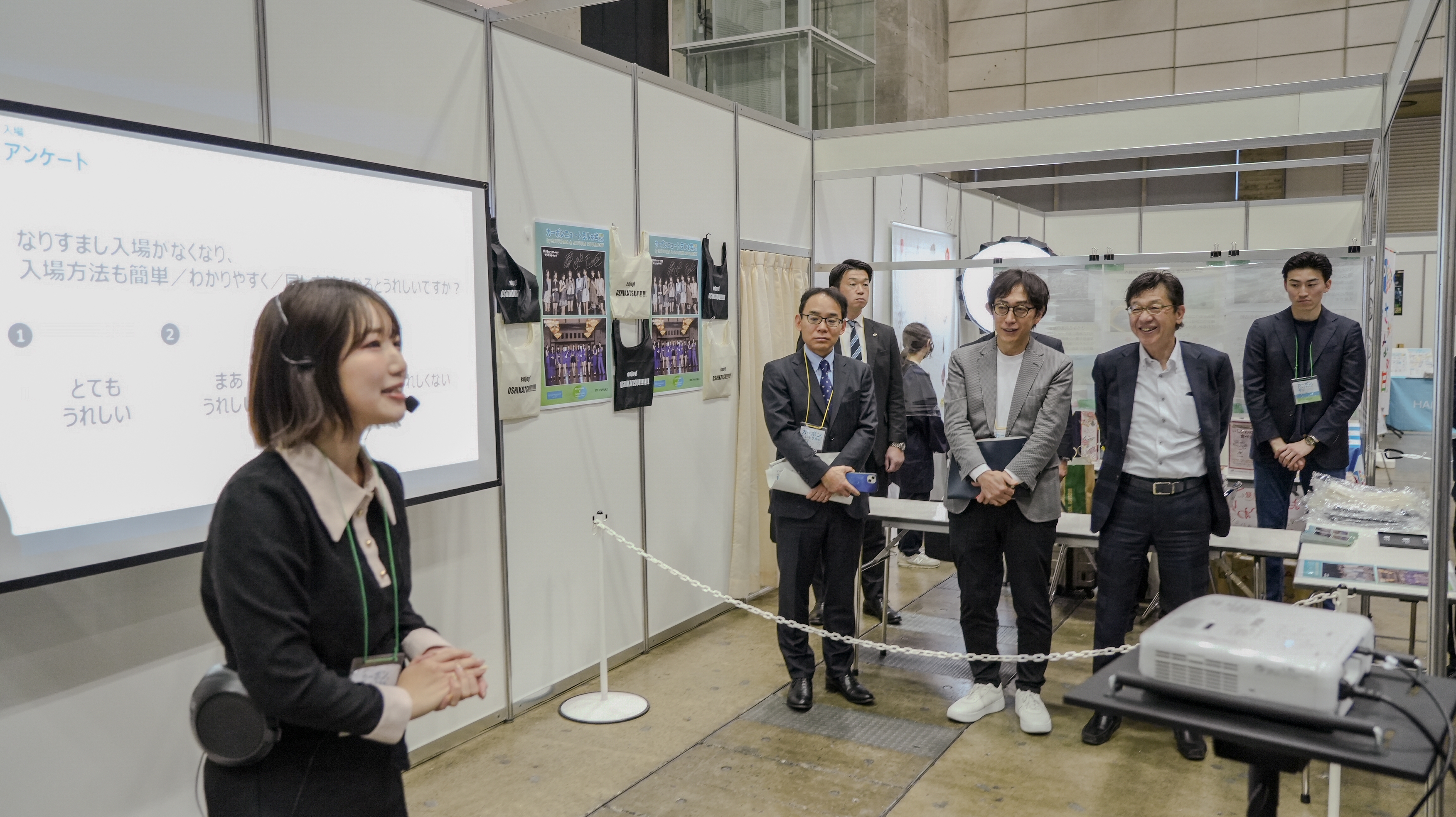 会場のブース内で、黒い服にヘッドセットマイクを着けた若い女性スタッフが説明を行っている。背後のスクリーンには「アンケート」と題したスライドが映し出されており、「なりすまし入場がなくなり、入場方法も簡単／わかりやすく／同じ…とうれしいですか？」という設問と、「①とてもうれしい」「②まあうれしい」などの選択肢が表示されている。ロープの内側にはスーツ姿の男性3名が並んで聴講している。ブース内にはグッズや写真パネルが展示されている。
